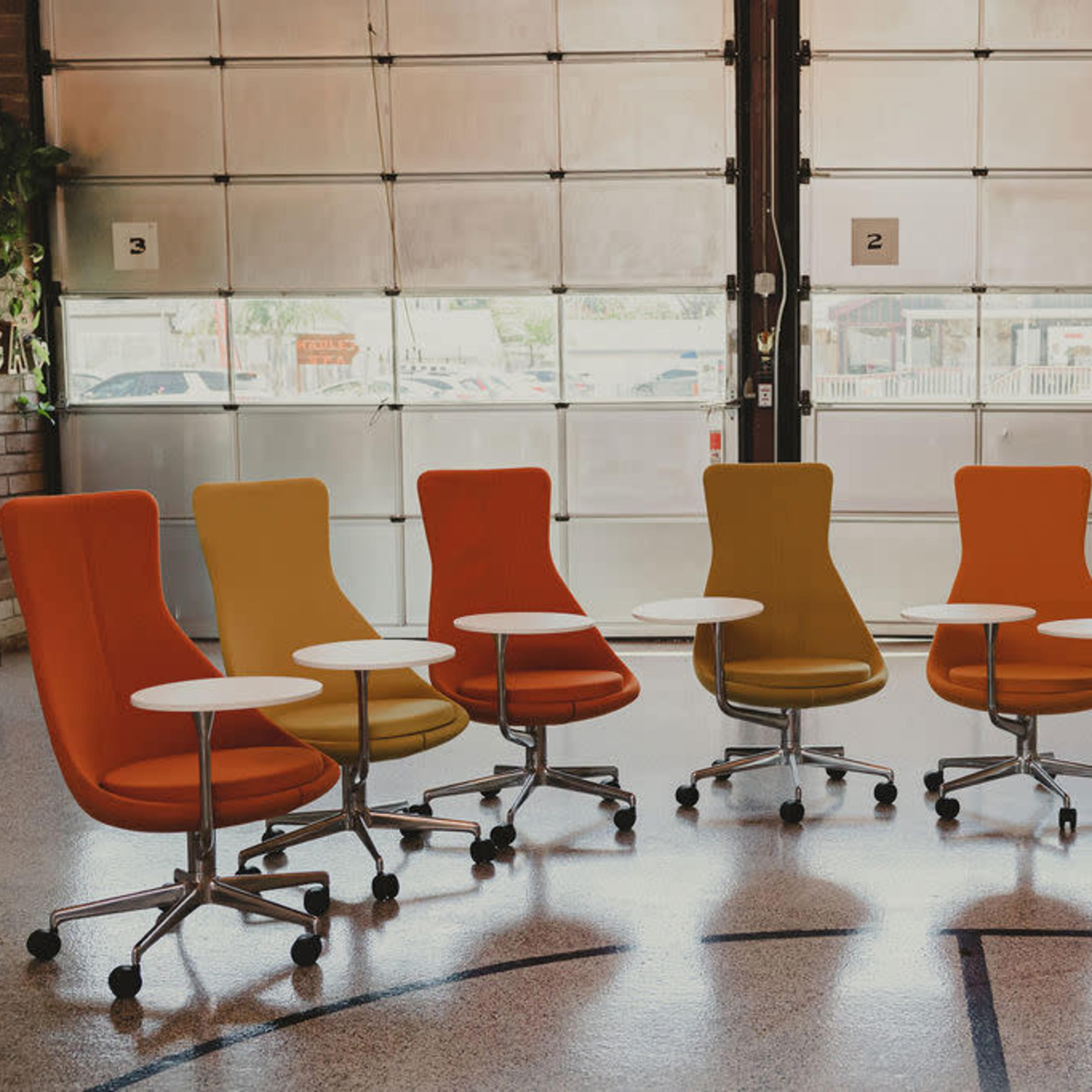 A group of five modern swivel chairs in yellow, orange, and gray is arranged around small white tables in a spacious room with large windows.