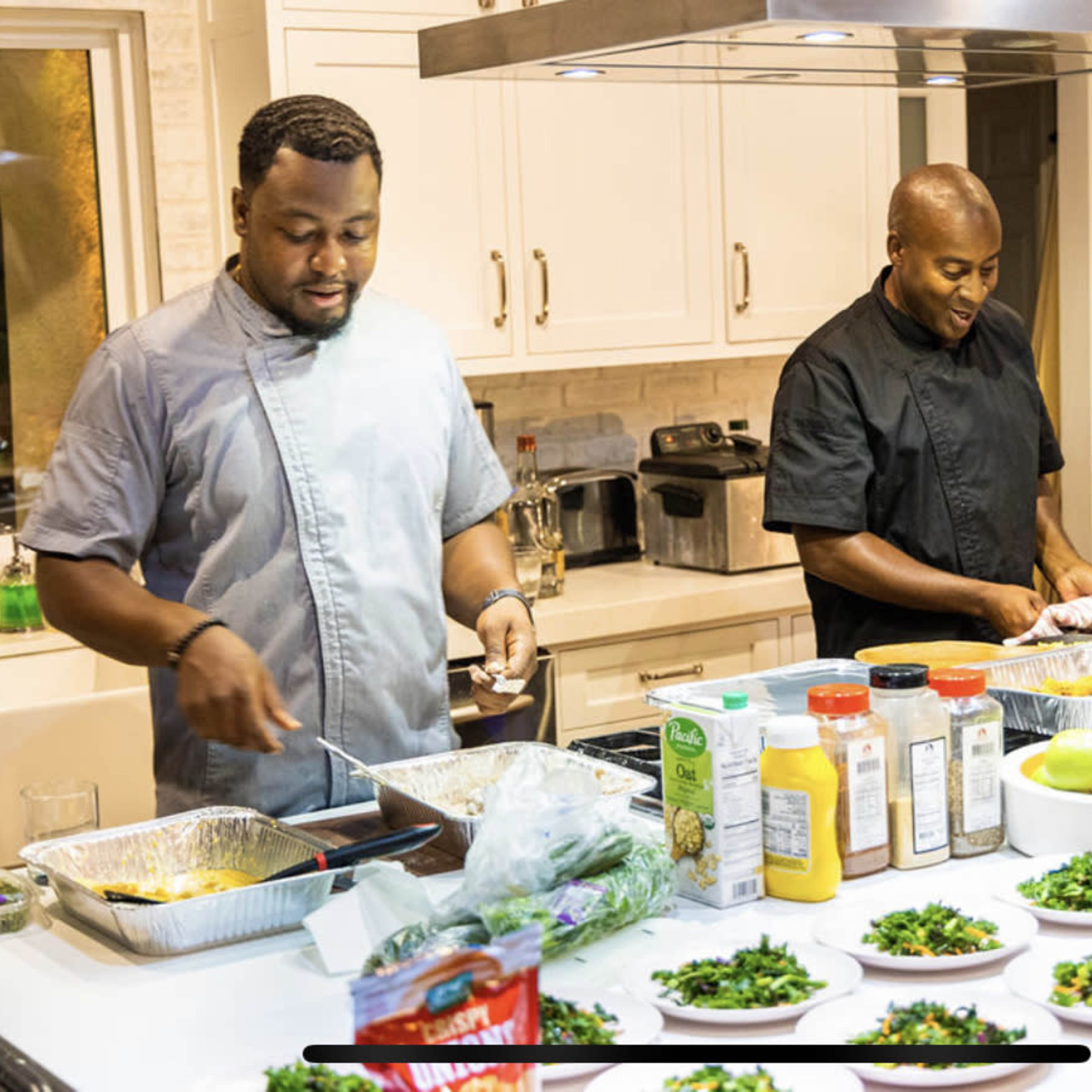 Two chefs are preparing food in a modern kitchen, with various dishes and ingredients arranged on the counter.
