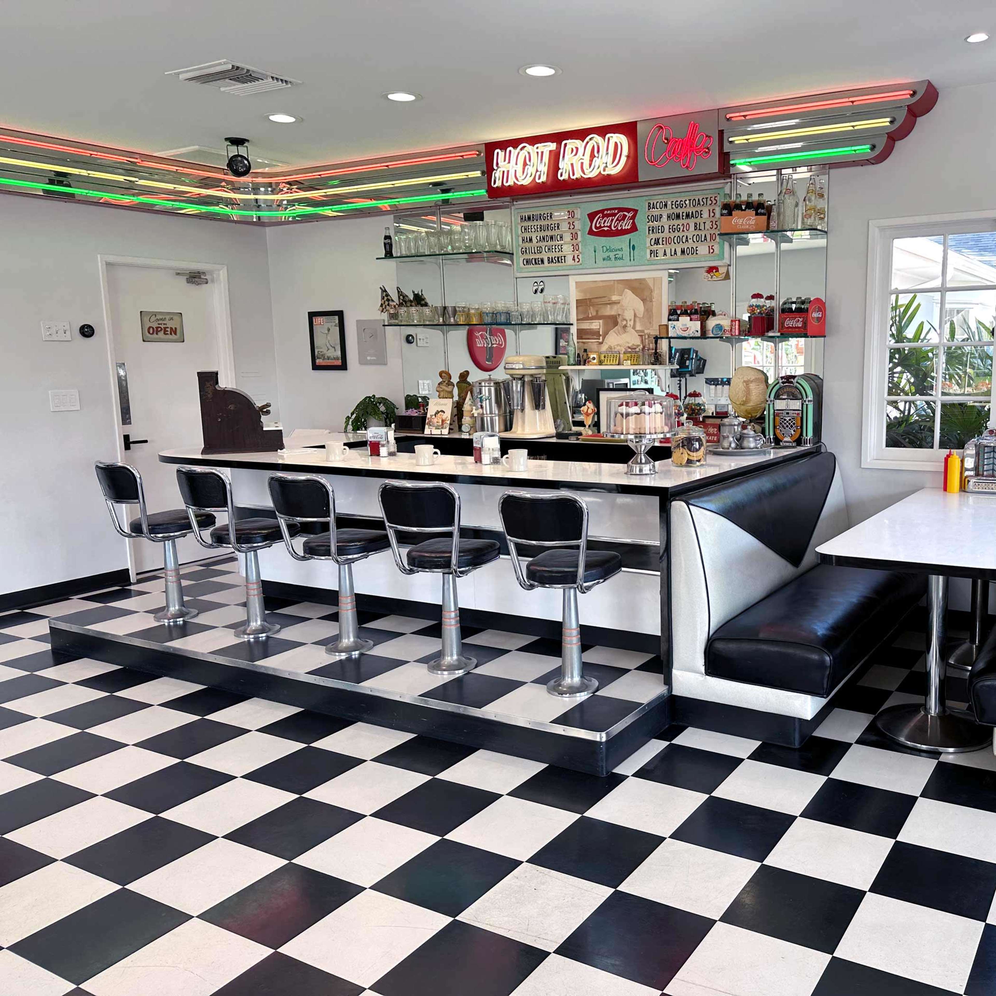 A retro diner with a checkered floor, a counter with barstools, and booths along the walls, featuring neon lighting and vintage decor.