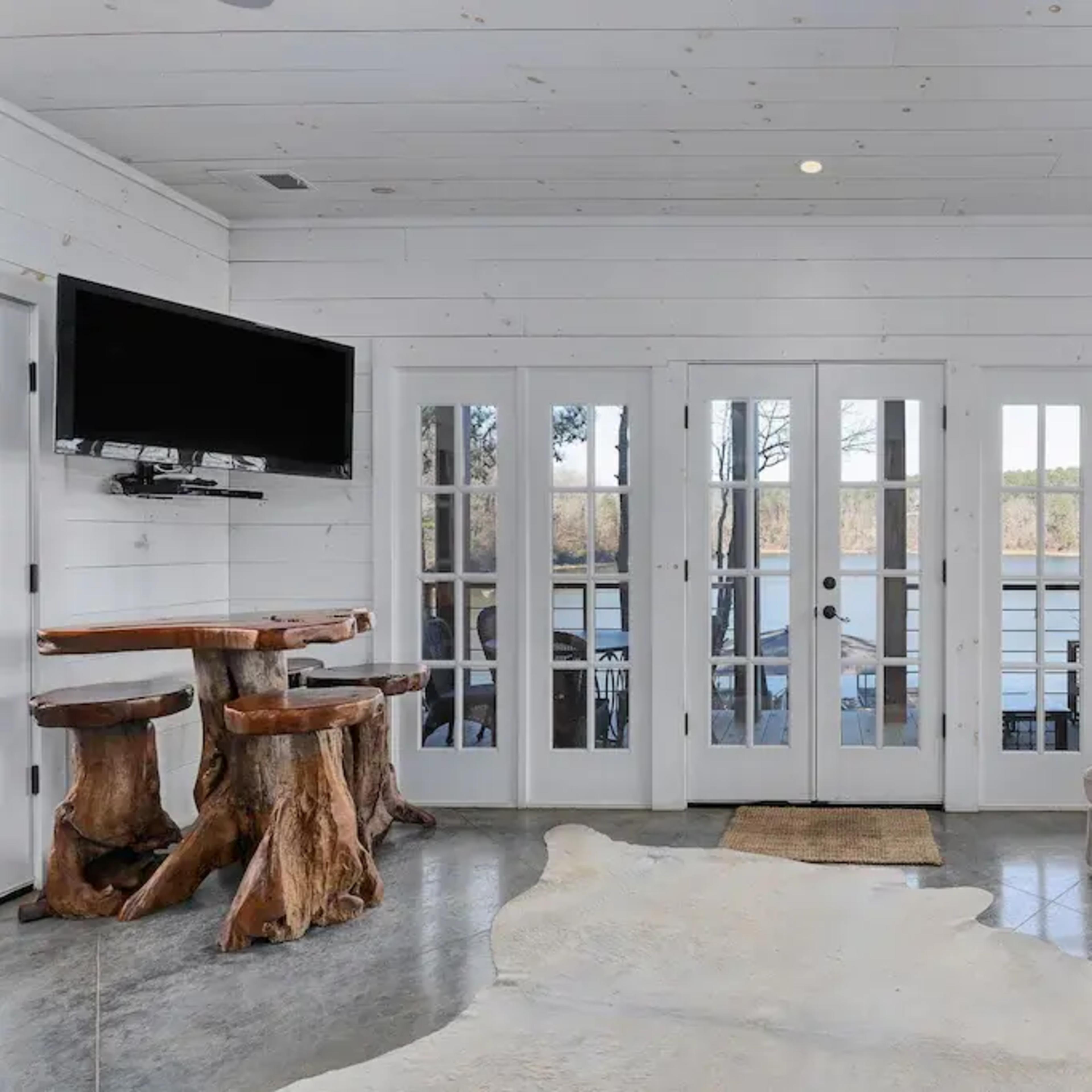 A room with white wooden walls, a seating area featuring a light-colored sofa, a wooden table made from tree stumps, and glass doors leading to an outdoor view.