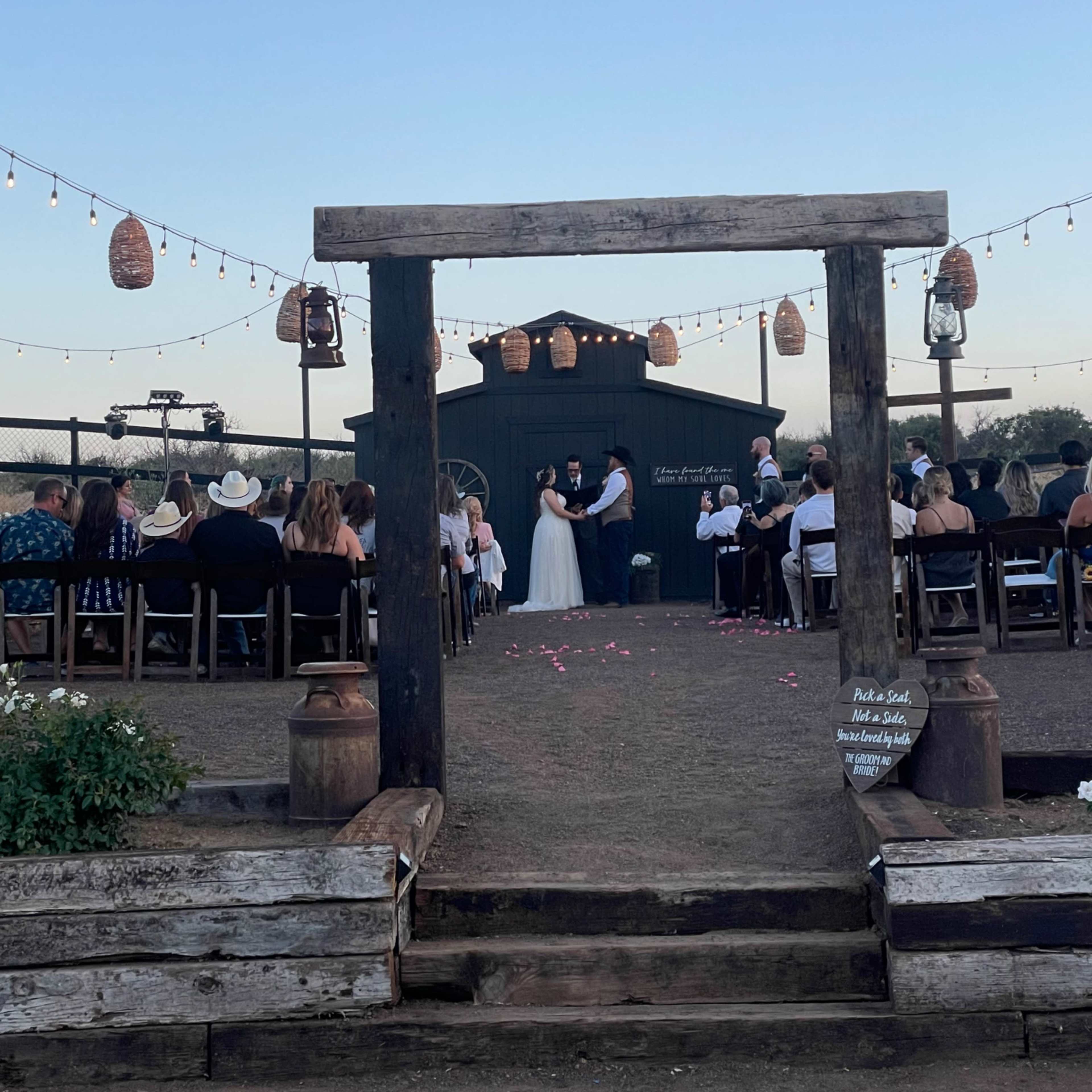 A couple exchanges vows under string lights and lanterns in a rustic outdoor wedding setting.