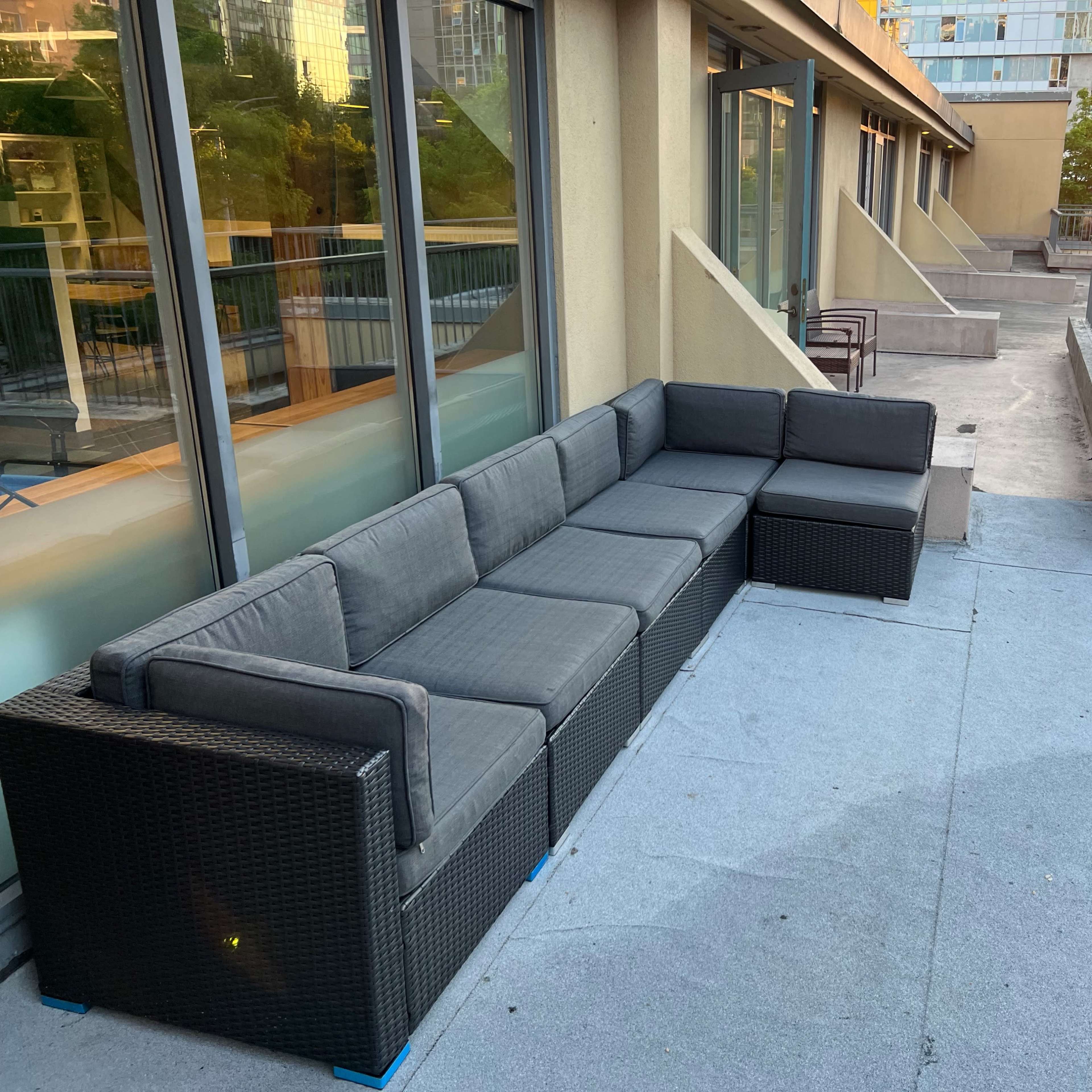 A large gray sectional sofa is positioned on a rooftop terrace adjacent to glass-walled buildings.