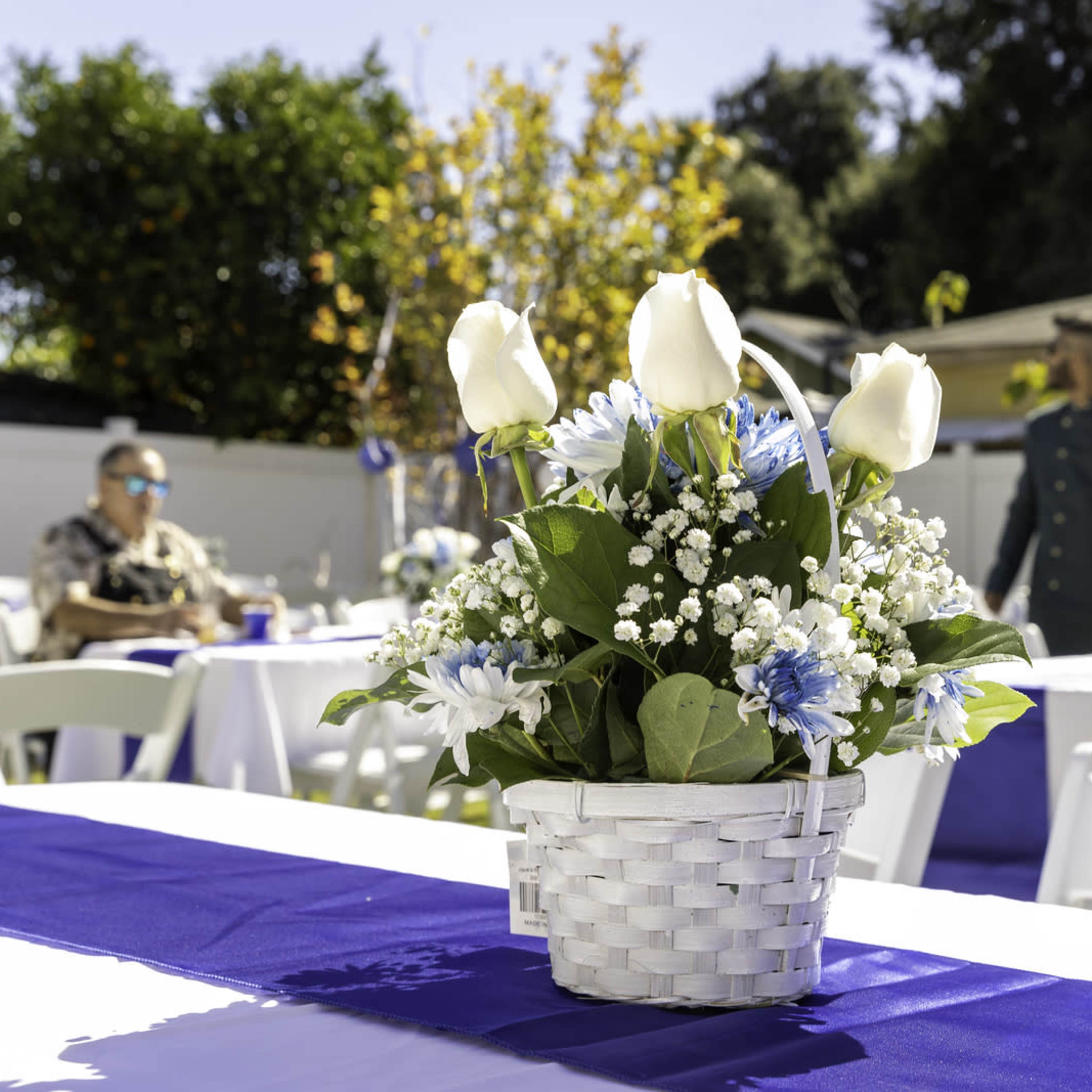 A centerpiece of white roses and blue flowers arranged in a woven basket sits on a table covered with a blue runner at an outdoor event.