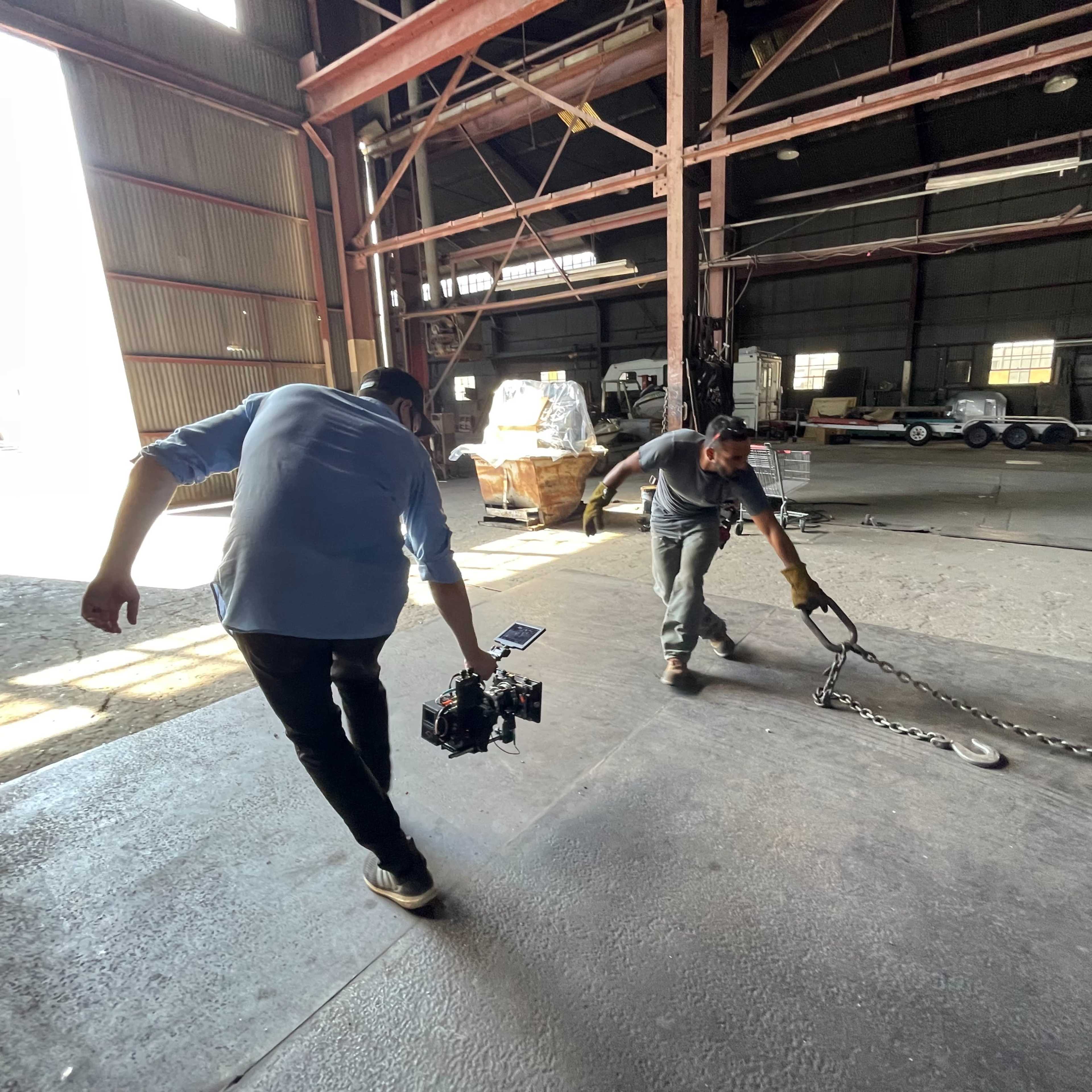 A man pulls a large metal chain on a workshop floor while another man carries a camera on a gimbal.