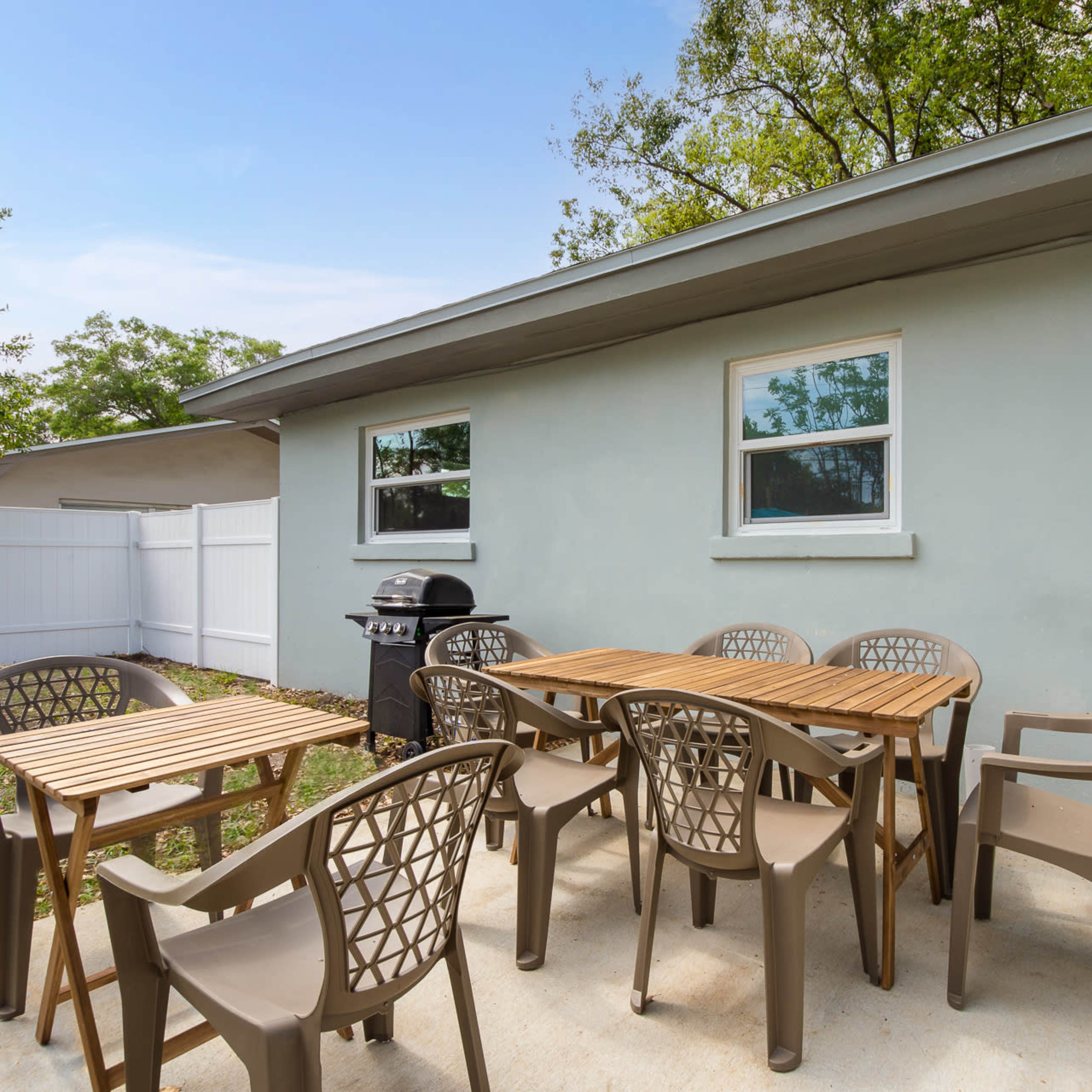 A patio features a wooden dining table surrounded by multiple chairs, with a grill and a fenced yard in the background.