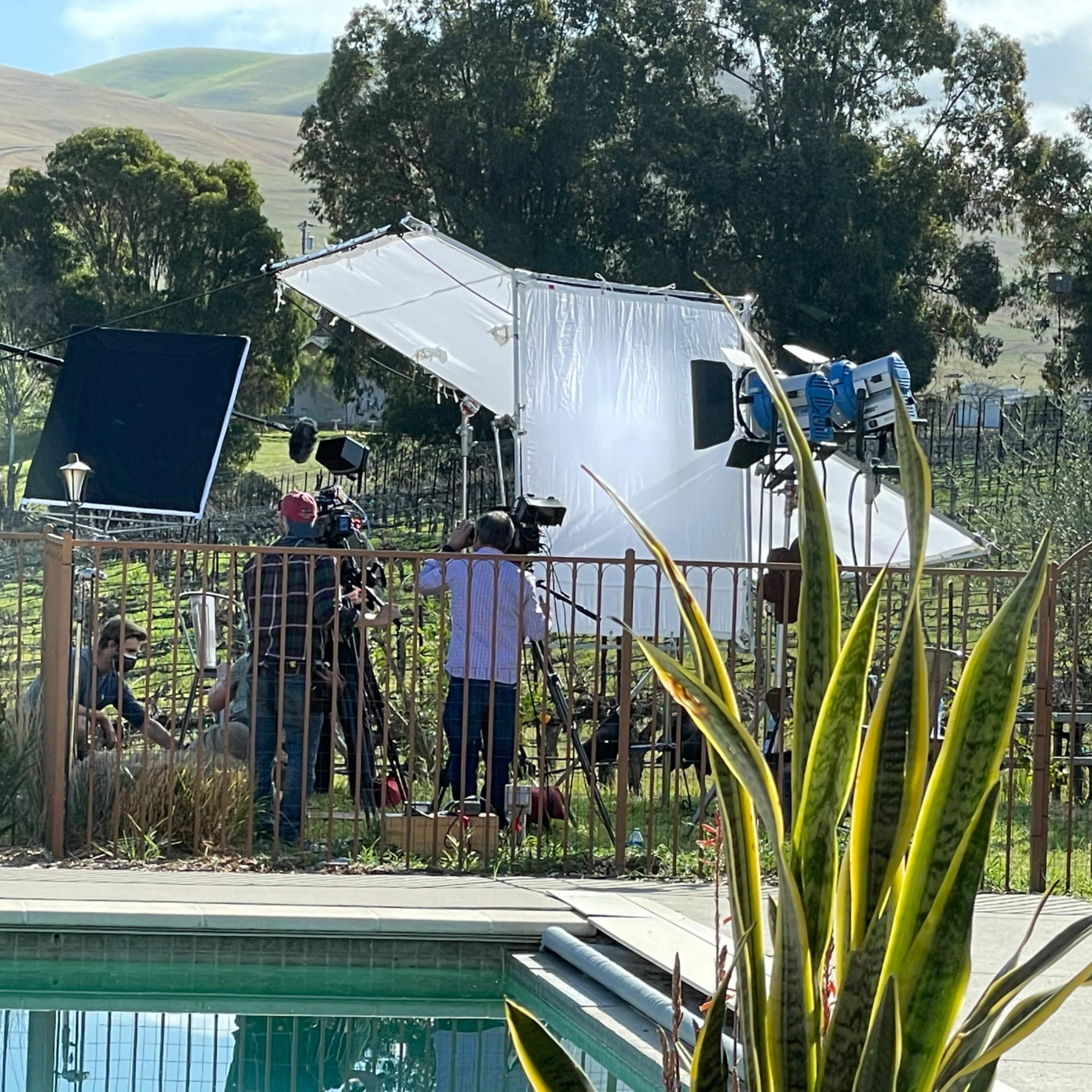 A film crew is set up near a swimming pool on a property with vineyards in the background, featuring large lighting equipment and cameras.