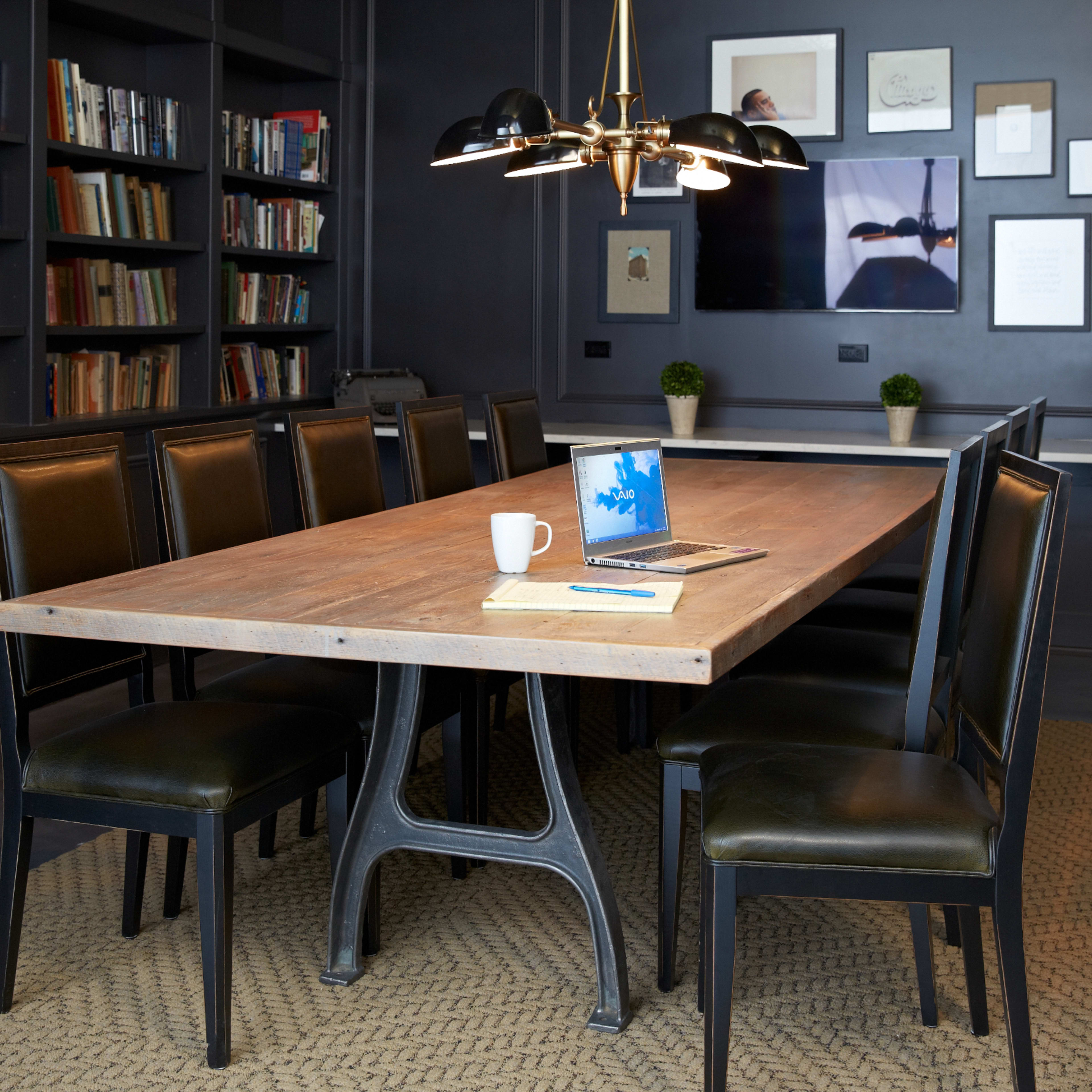 A long wooden table with eight black chairs is set in a room with bookshelves and a large wall-mounted screen.
