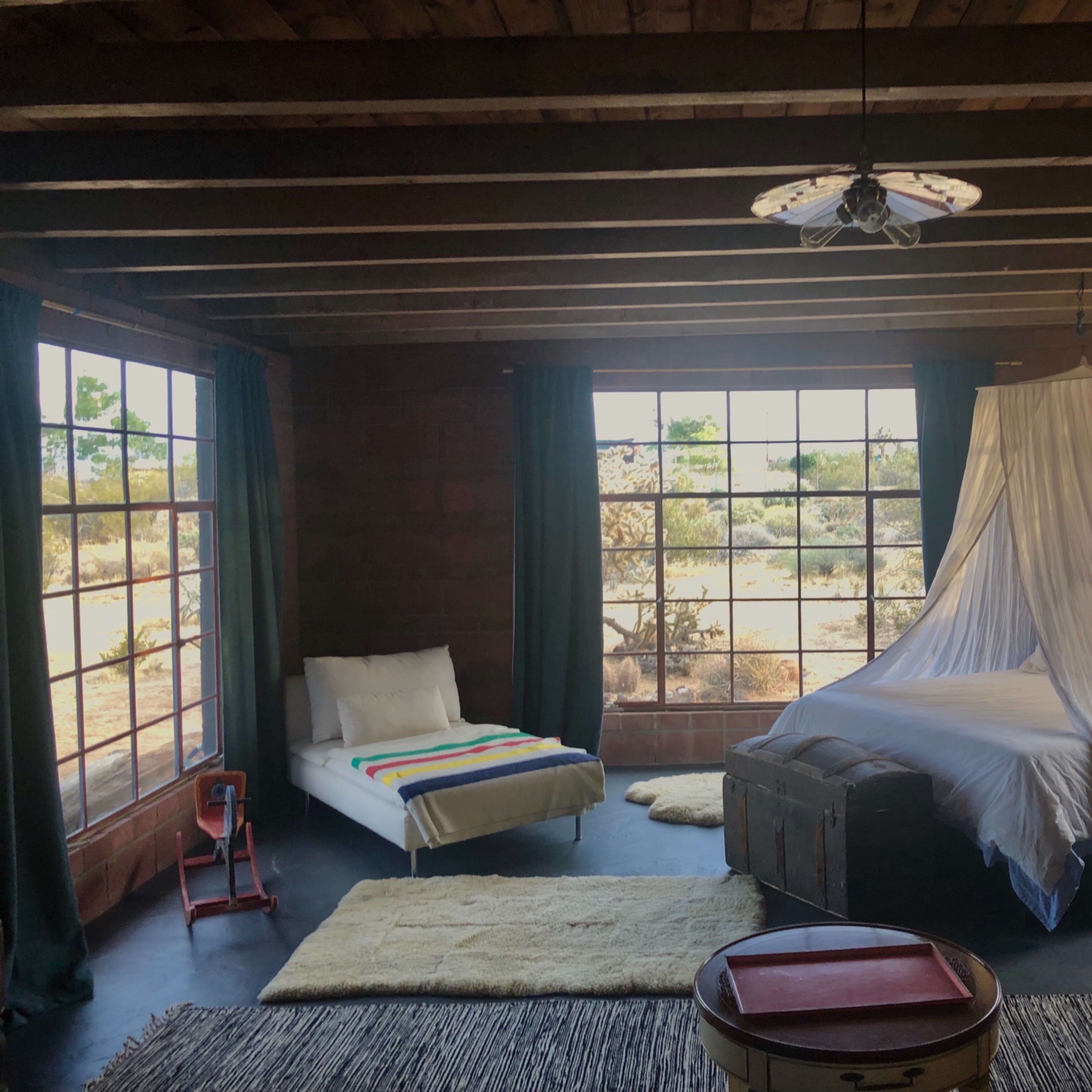 A rustic bedroom features a bed with a canopy, a white lounge chair, and large windows overlooking a desert landscape.
