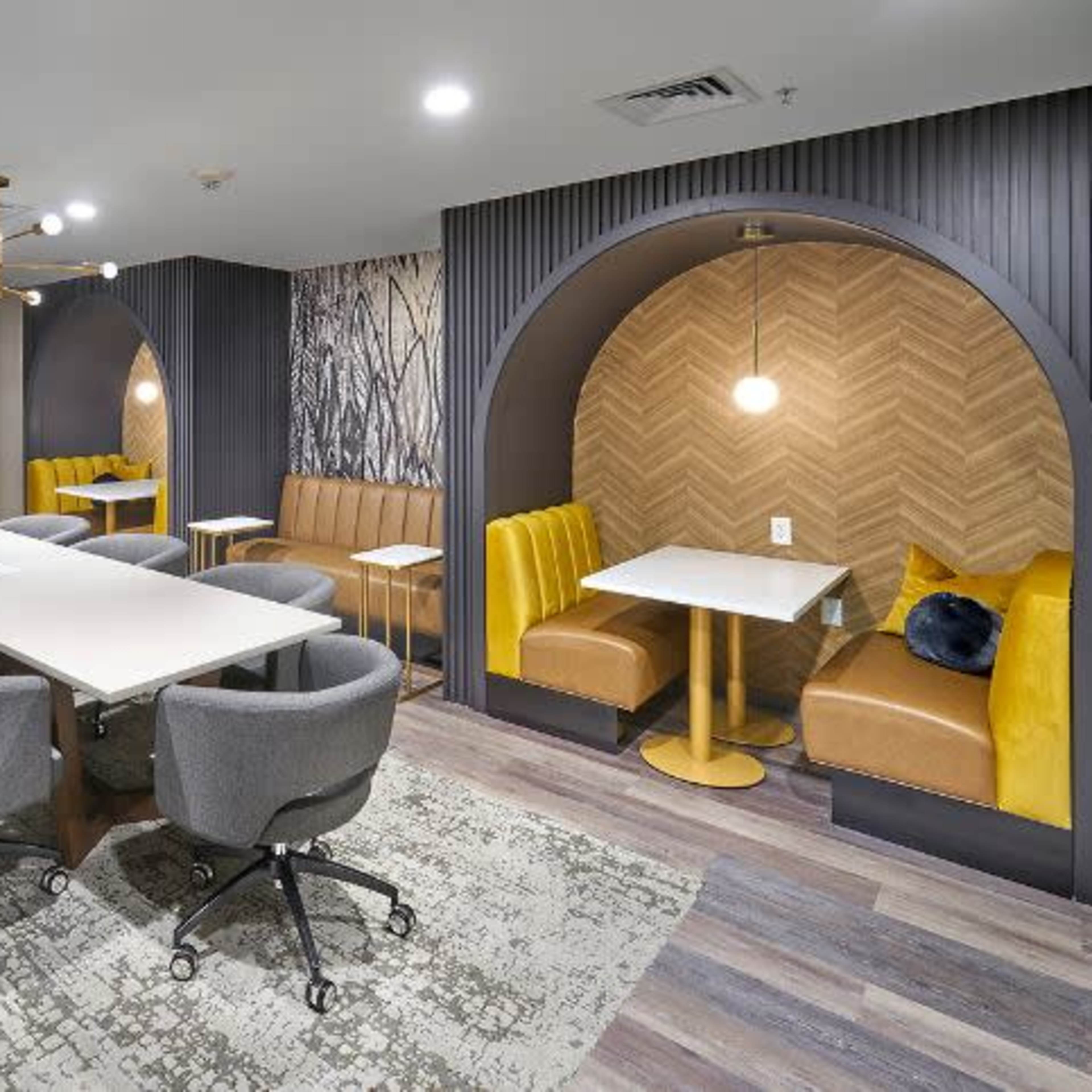 A modern meeting room with a long table surrounded by chairs, two booth seating areas, and a wall-mounted television.