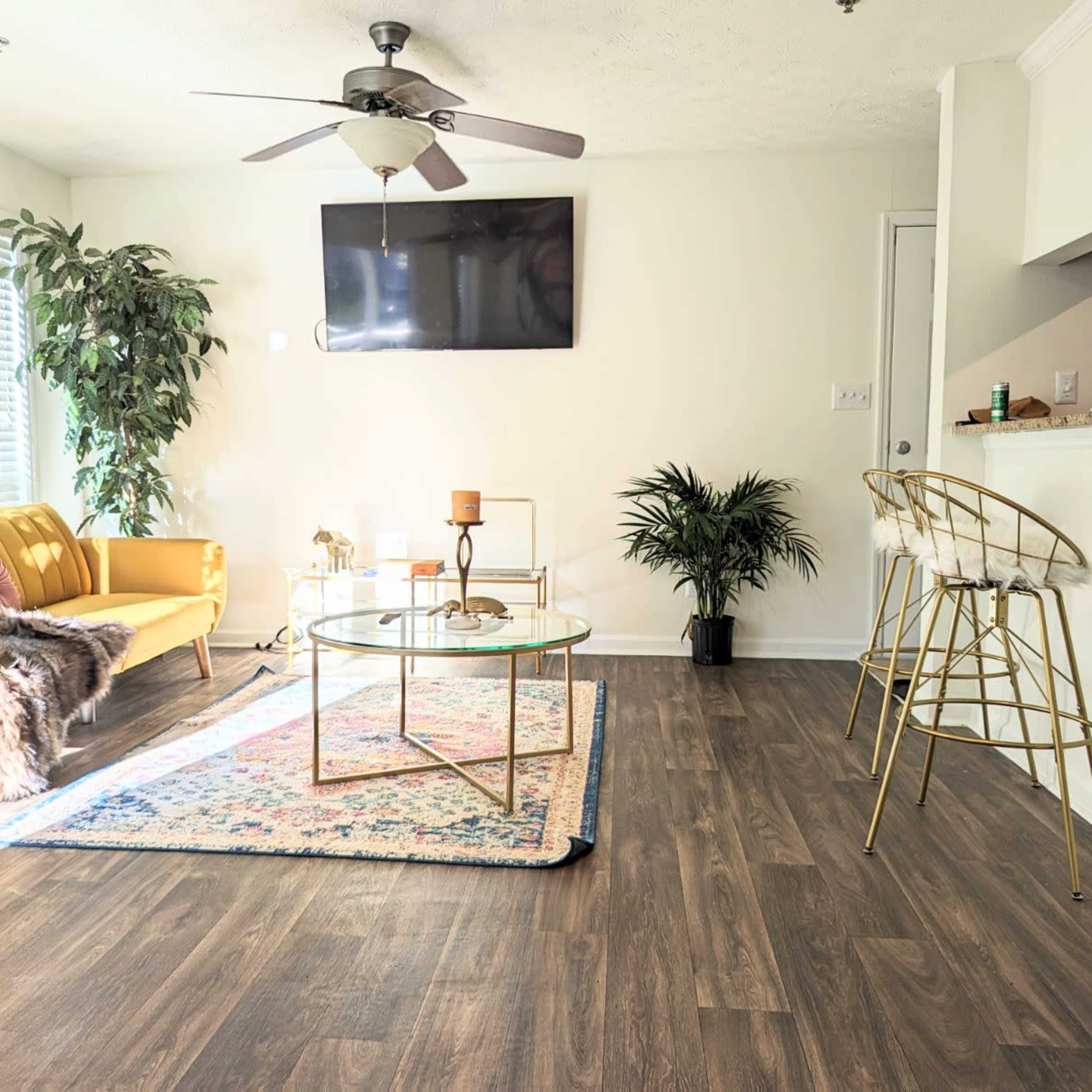 A bright living room featuring a yellow sofa, a glass coffee table on a colorful rug, and bar stools near a kitchen counter.