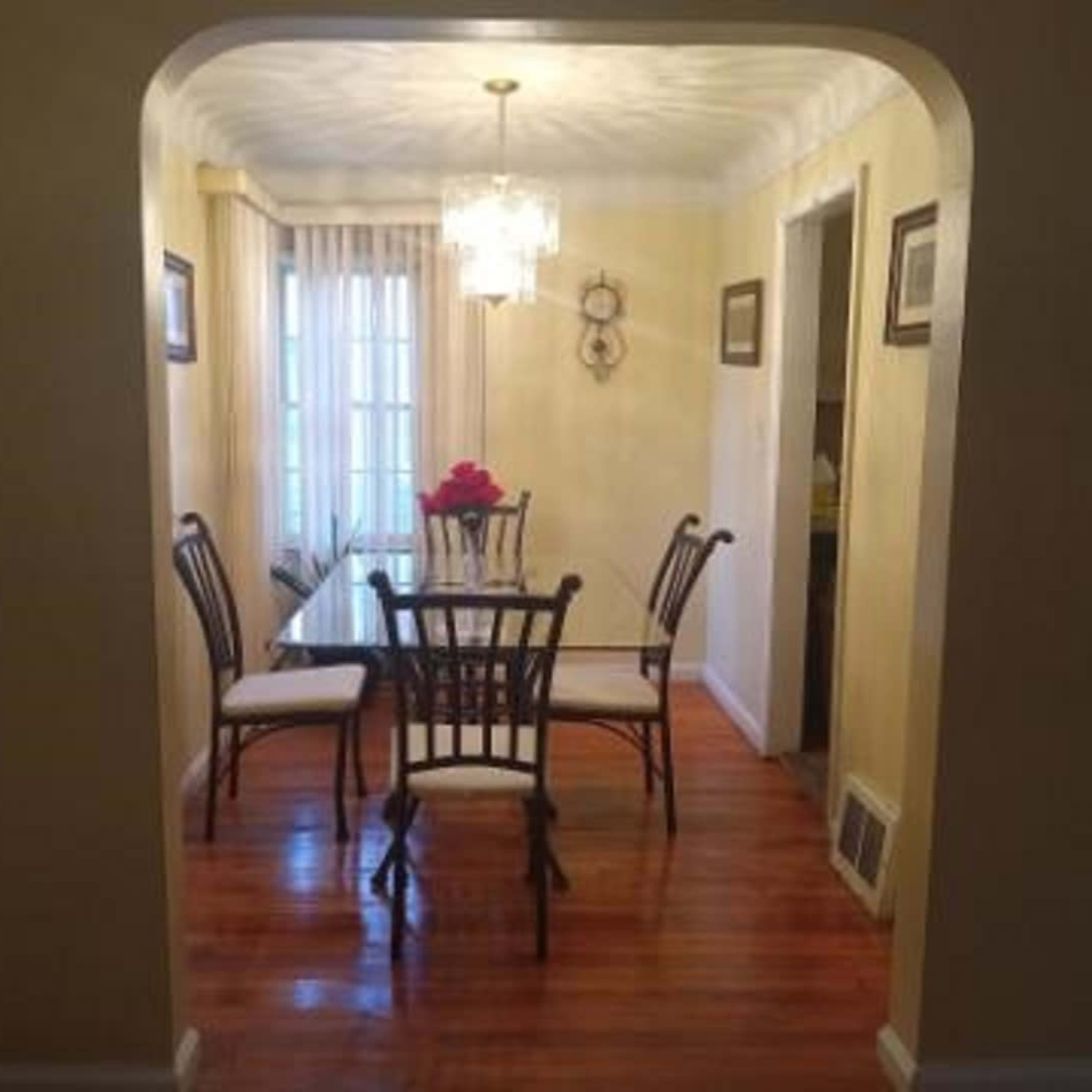A dining area features a wooden table surrounded by six chairs, adorned with a vase of flowers, and illuminated by a hanging chandelier.
