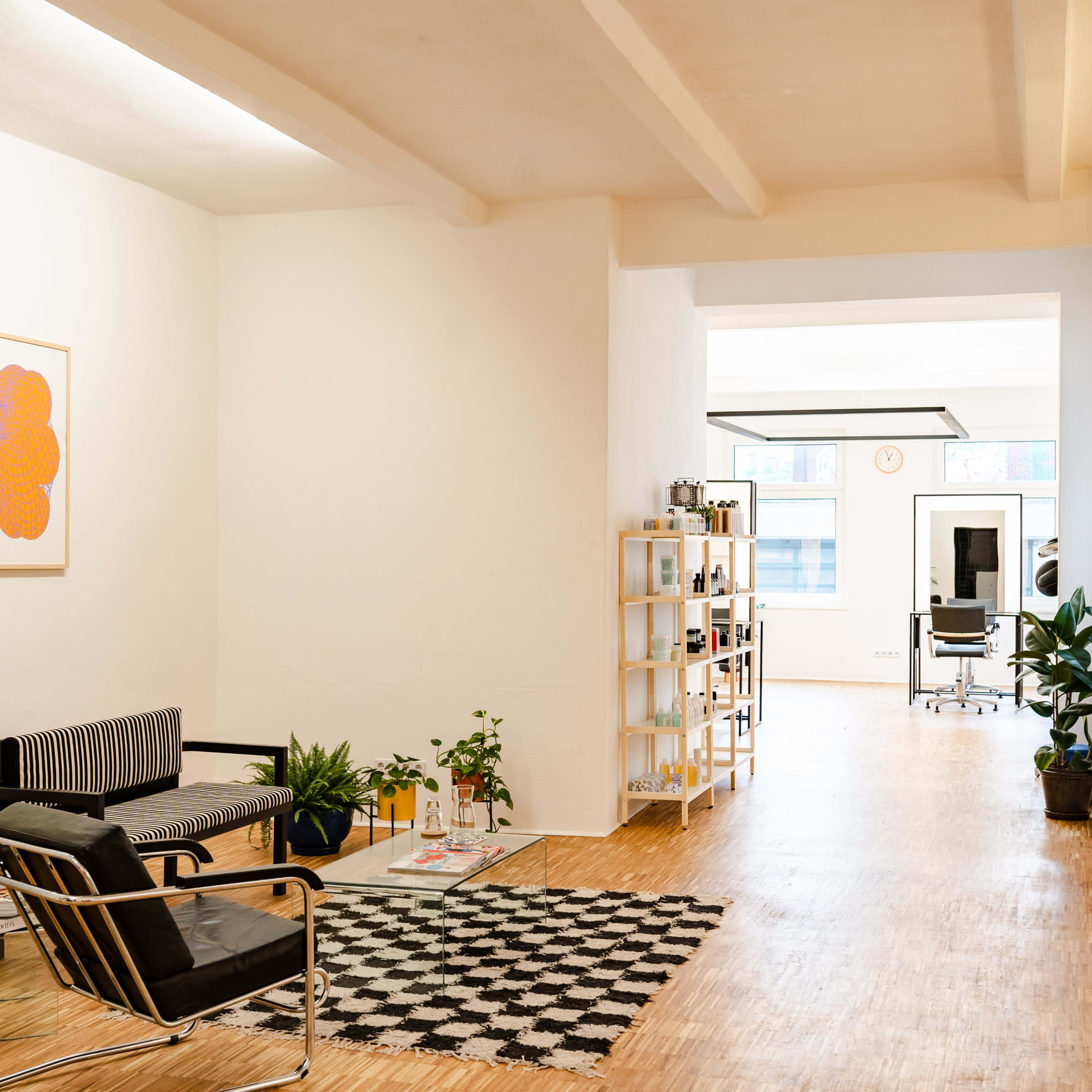 A spacious, modern office area with light wooden floors, plants, and a mix of seating and shelving arrangements.
