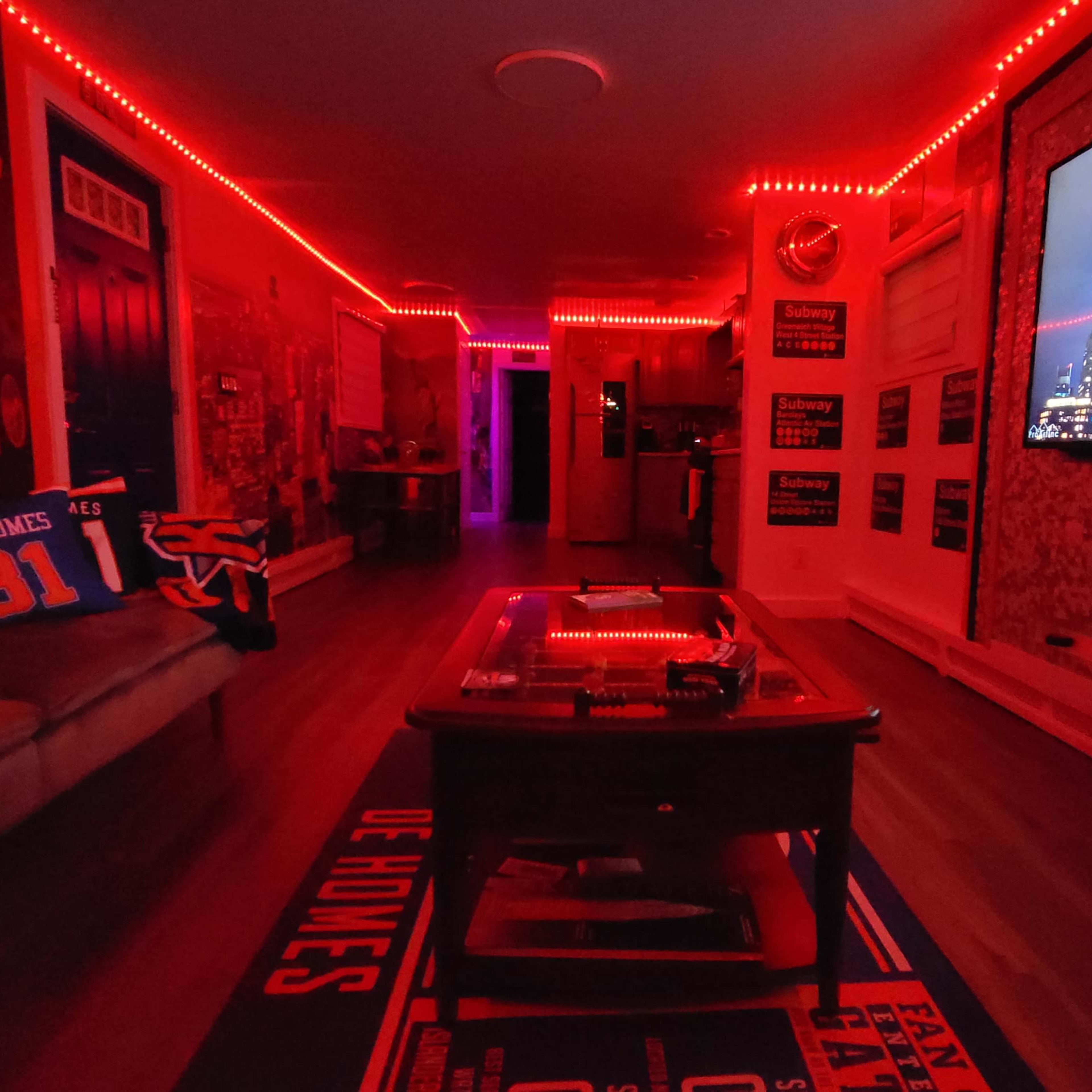 The room is illuminated with red LED lights, featuring a large screen displaying city skyline images, a couch with themed pillows, and a coffee table.