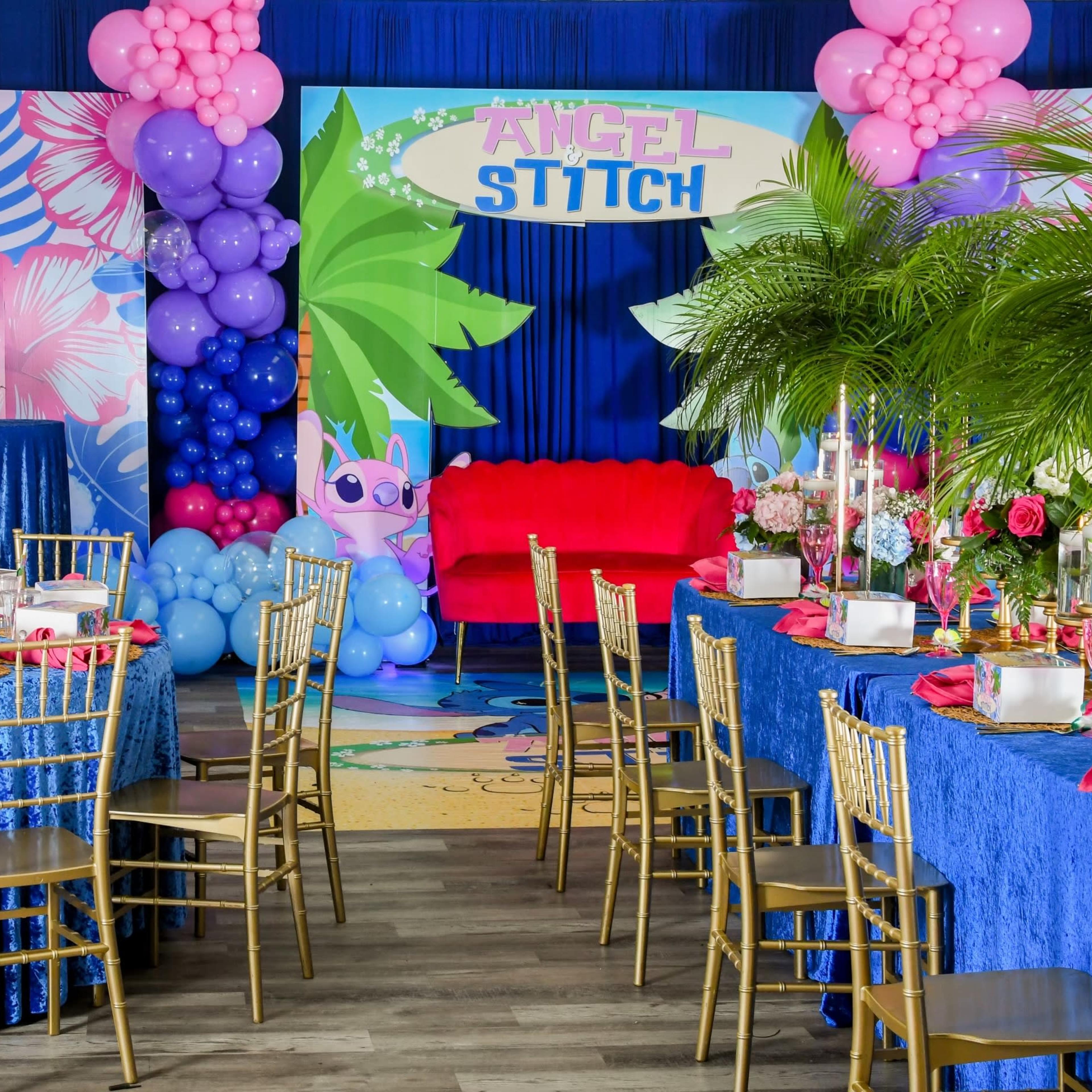 A party setup featuring two round tables with blue tablecloths, gold chairs, colorful balloon decorations, and a backdrop with the words "Angel Stitch" along with tropical themed elements.