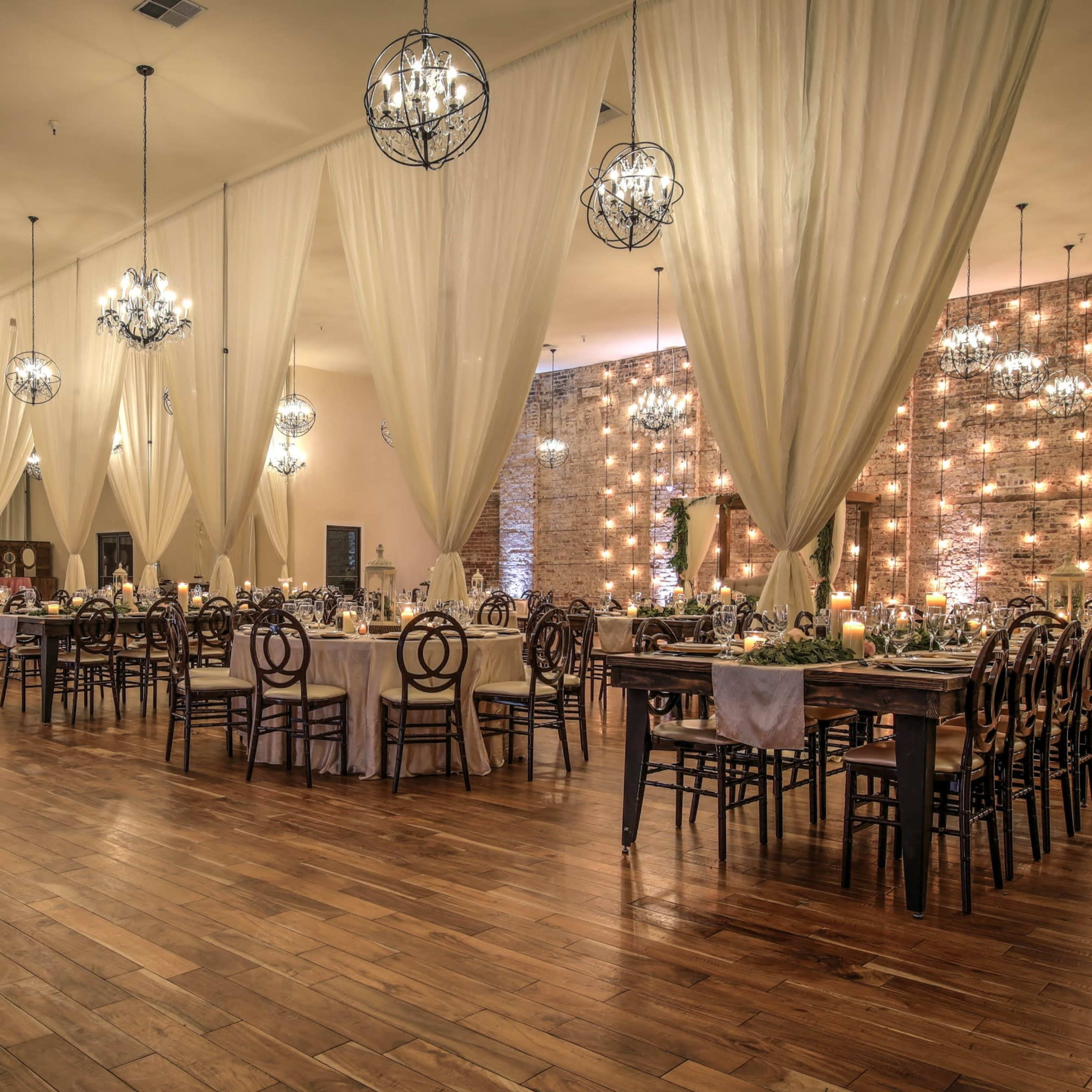 A spacious event hall featuring wooden floors, long dining tables, and chandeliers, with draped fabric along the ceiling and a brick wall adorned with lights.