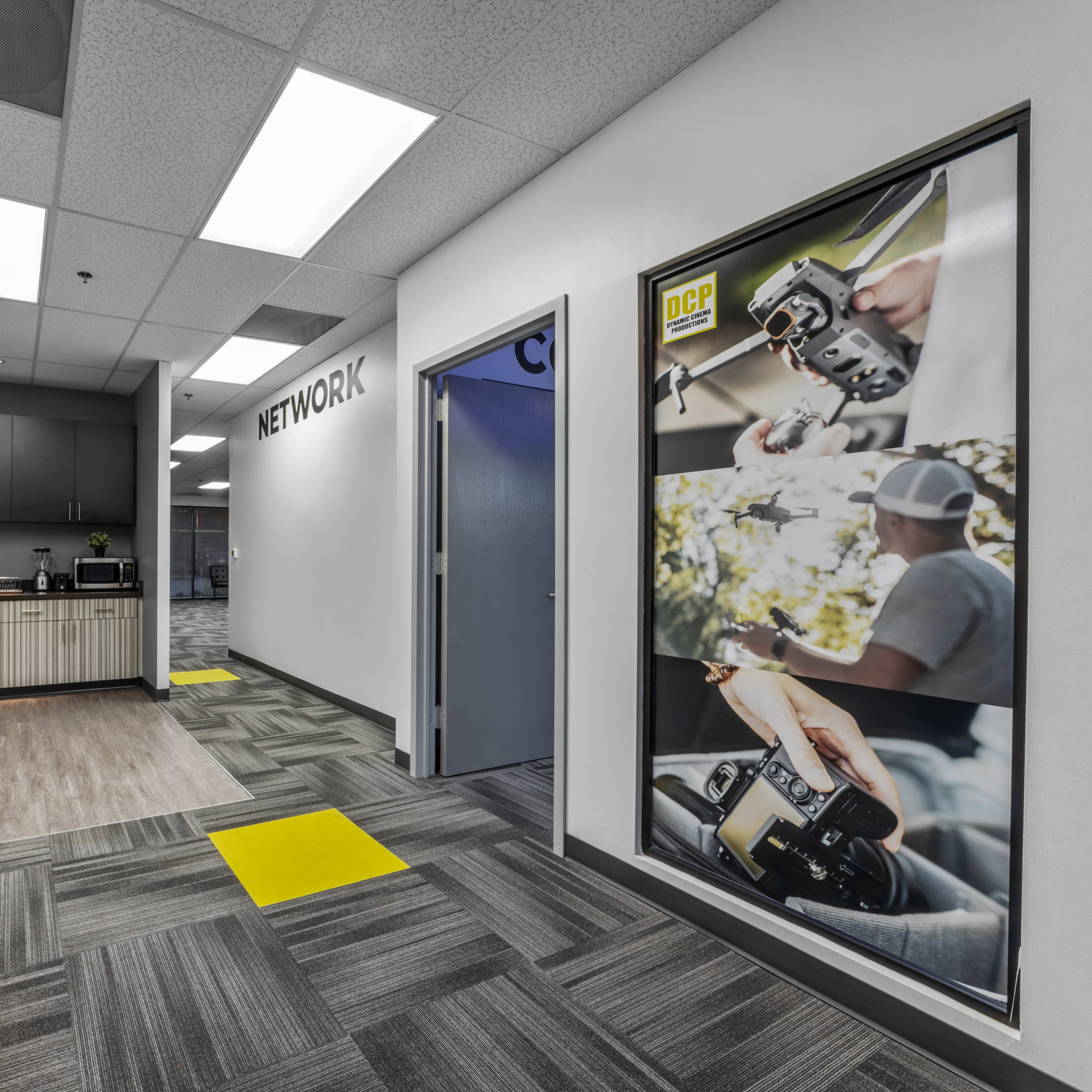 A modern office hallway features a kitchen area on one side and a large poster of a photographer on the other, with yellow and gray floor tiles.