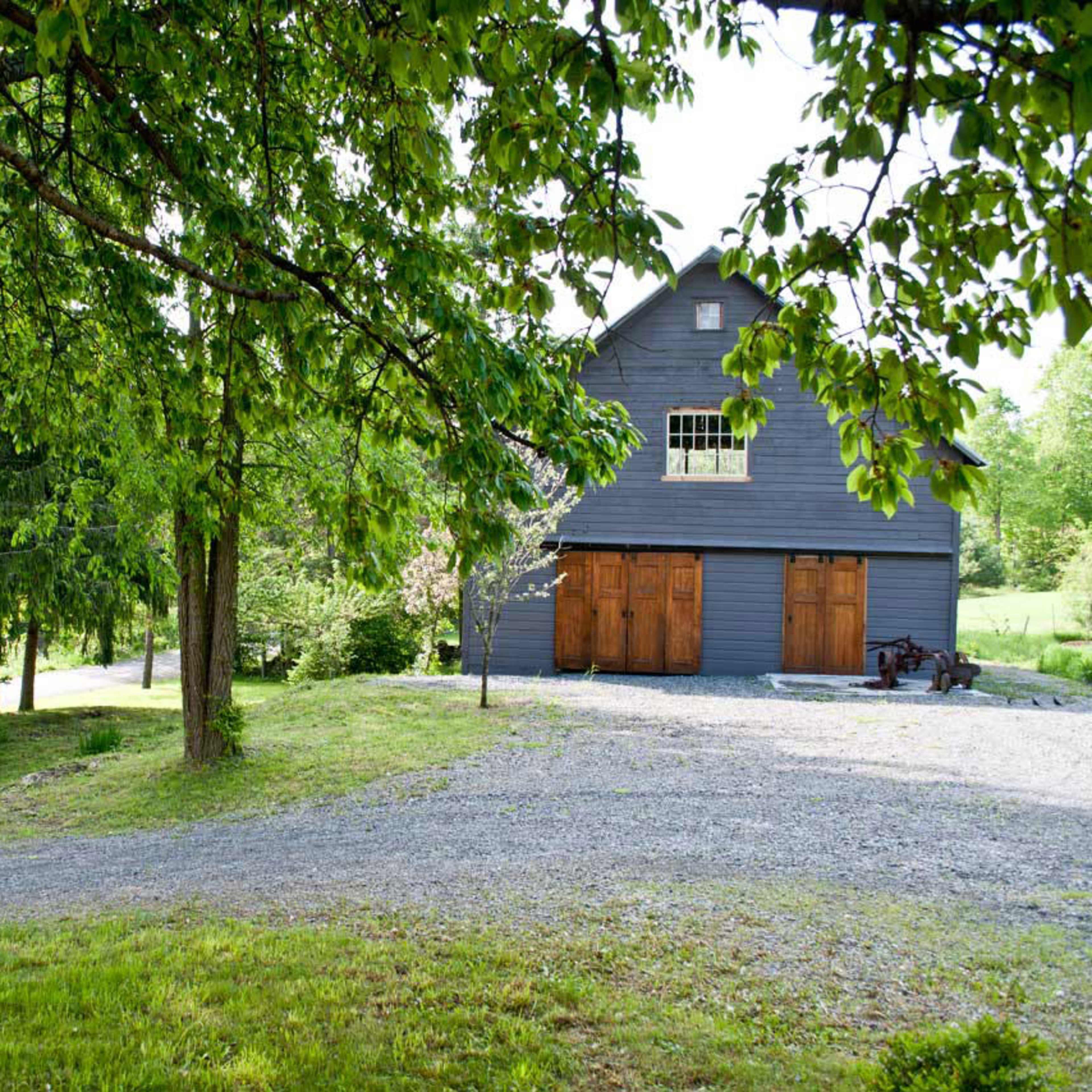 A large gray barn with wooden doors stands at the end of a gravel driveway surrounded by trees.