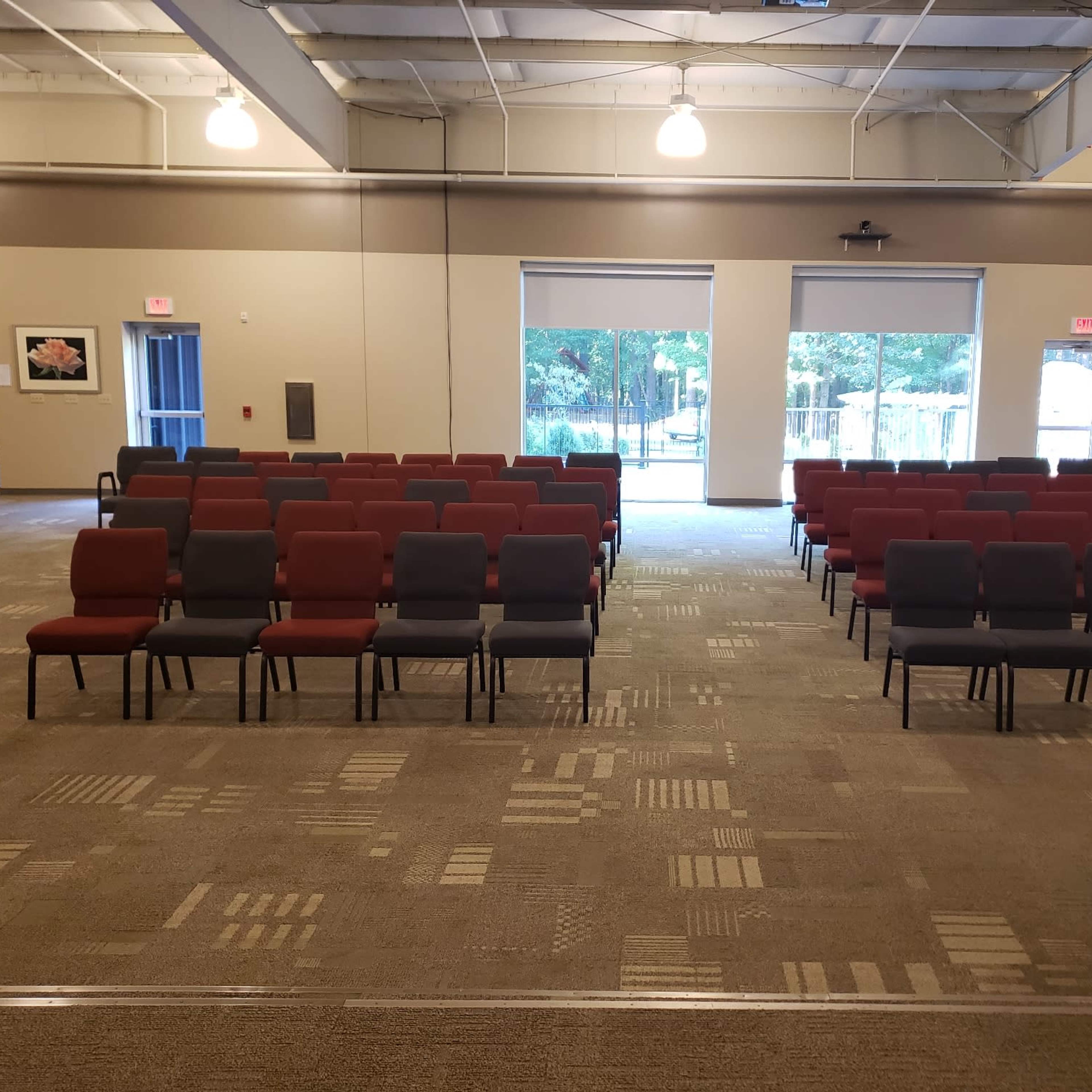 A large room arranged with rows of chairs, alternating between red and gray, facing a window with a view of greenery outside.