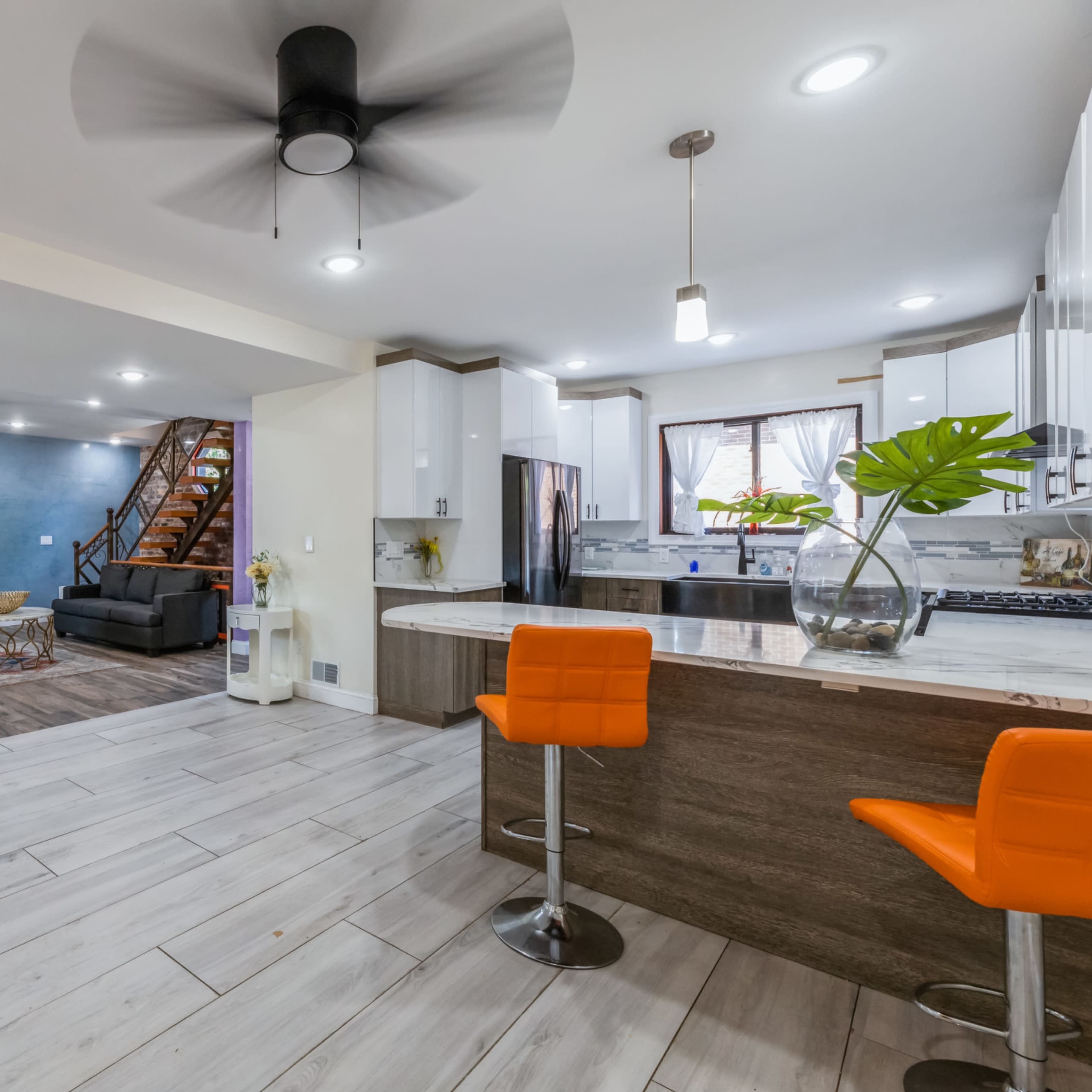 A modern open-concept living space with a kitchen featuring a gray and white color scheme, a dining area with orange bar stools, and a living area that includes a black sofa and a media unit.