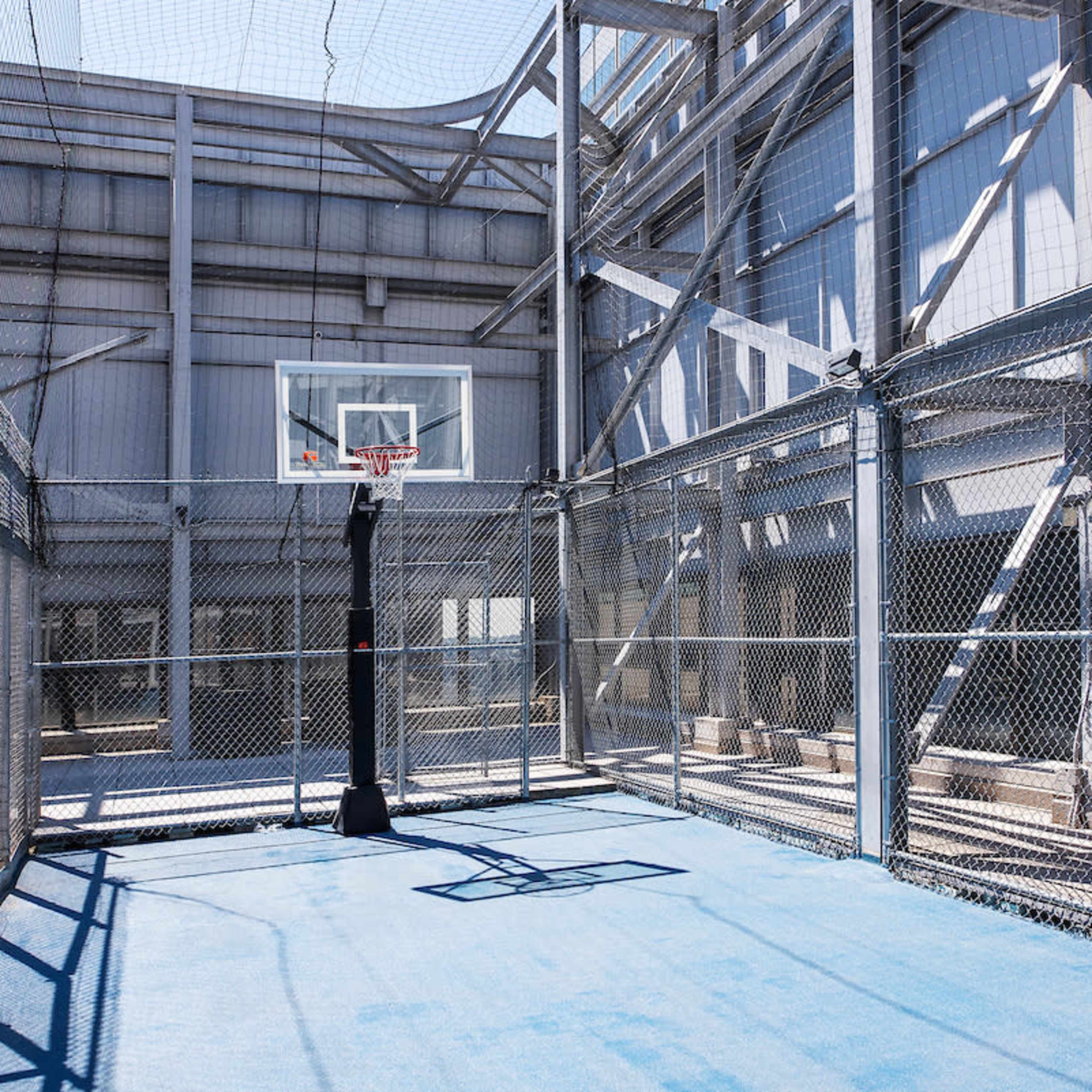 A fenced basketball court with a hoop and blue flooring, situated in a partially enclosed industrial area.