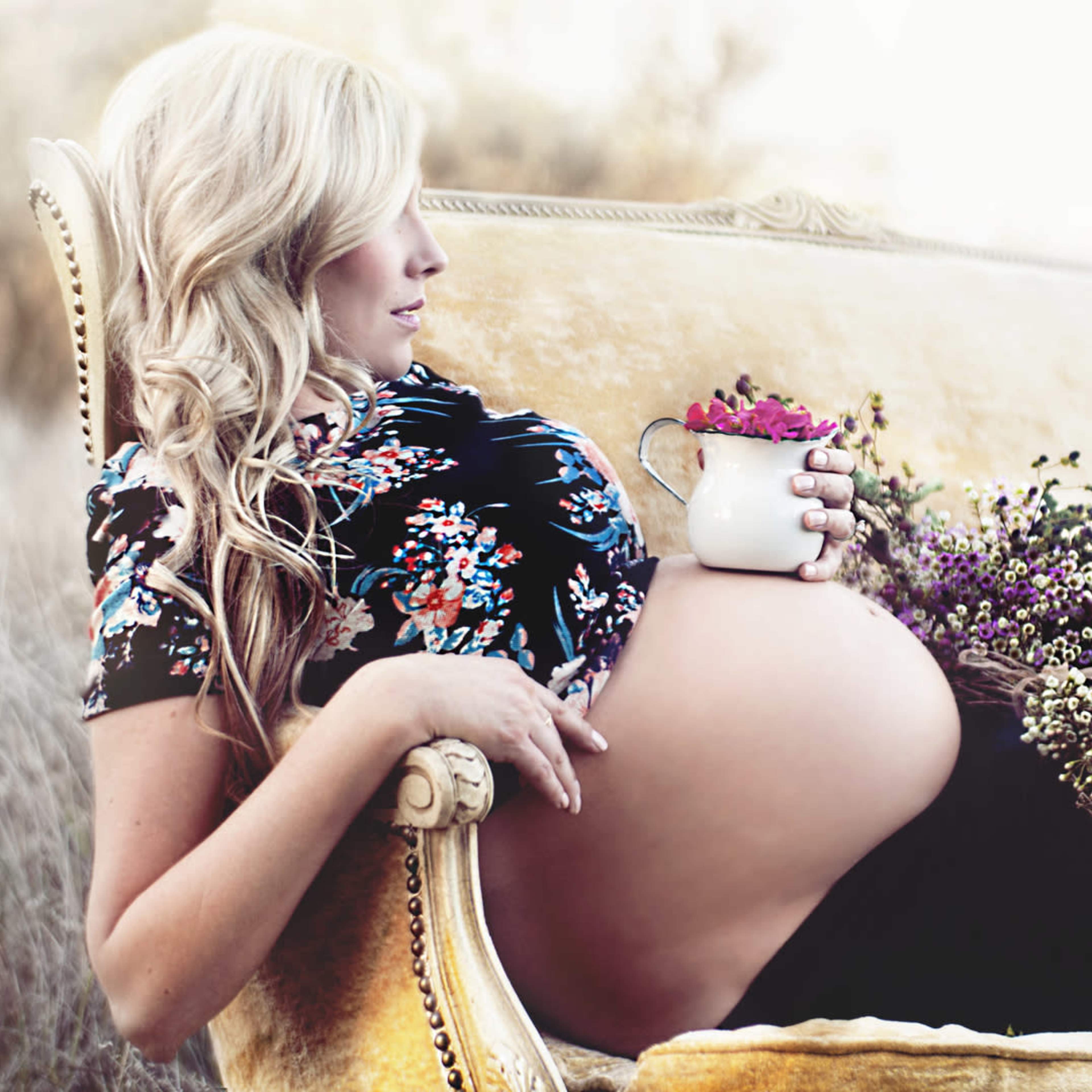 A pregnant woman sits on a vintage couch, holding a small vase of flowers and a larger bouquet beside her.