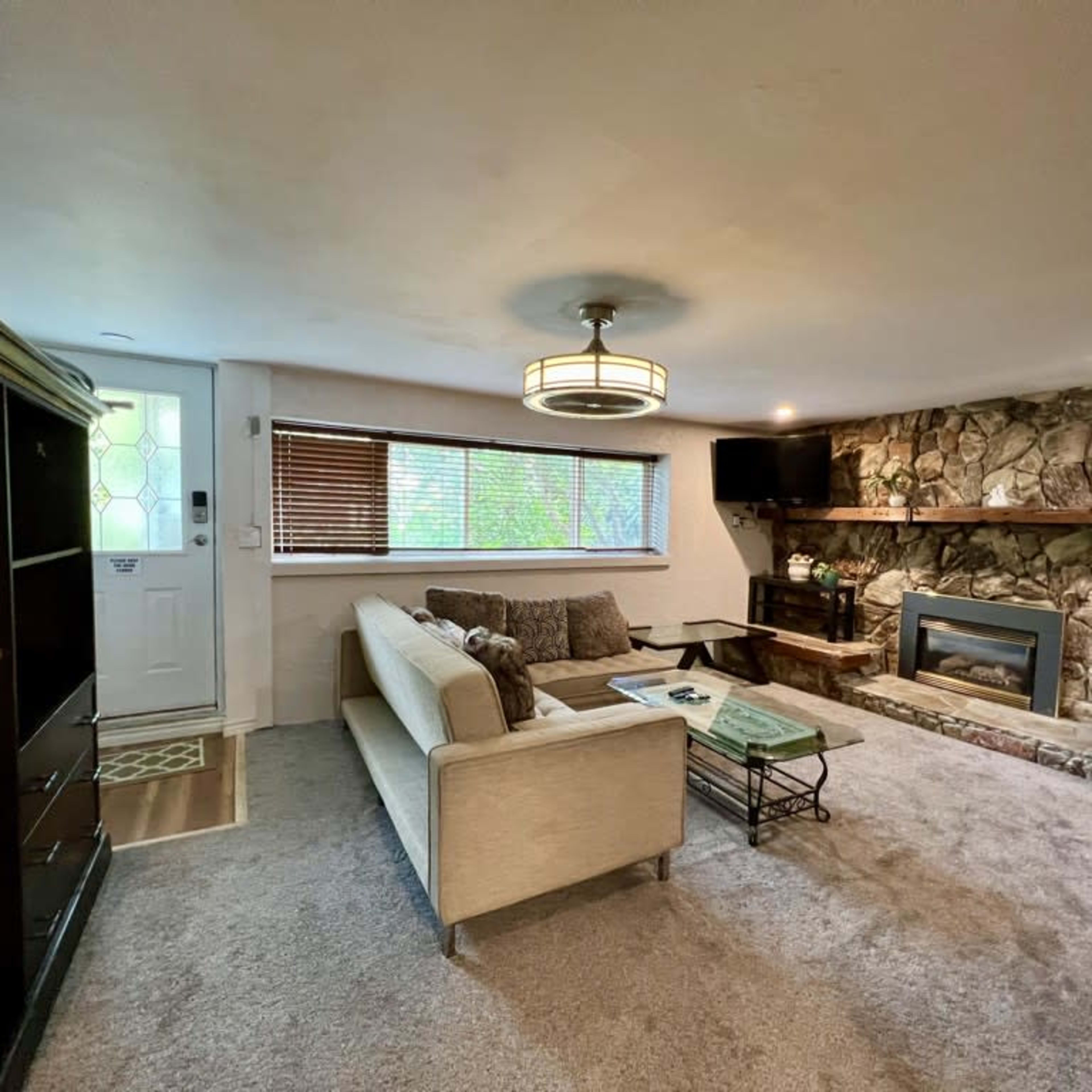 The living room features a beige sofa, a stone fireplace, and a large window with wooden blinds.