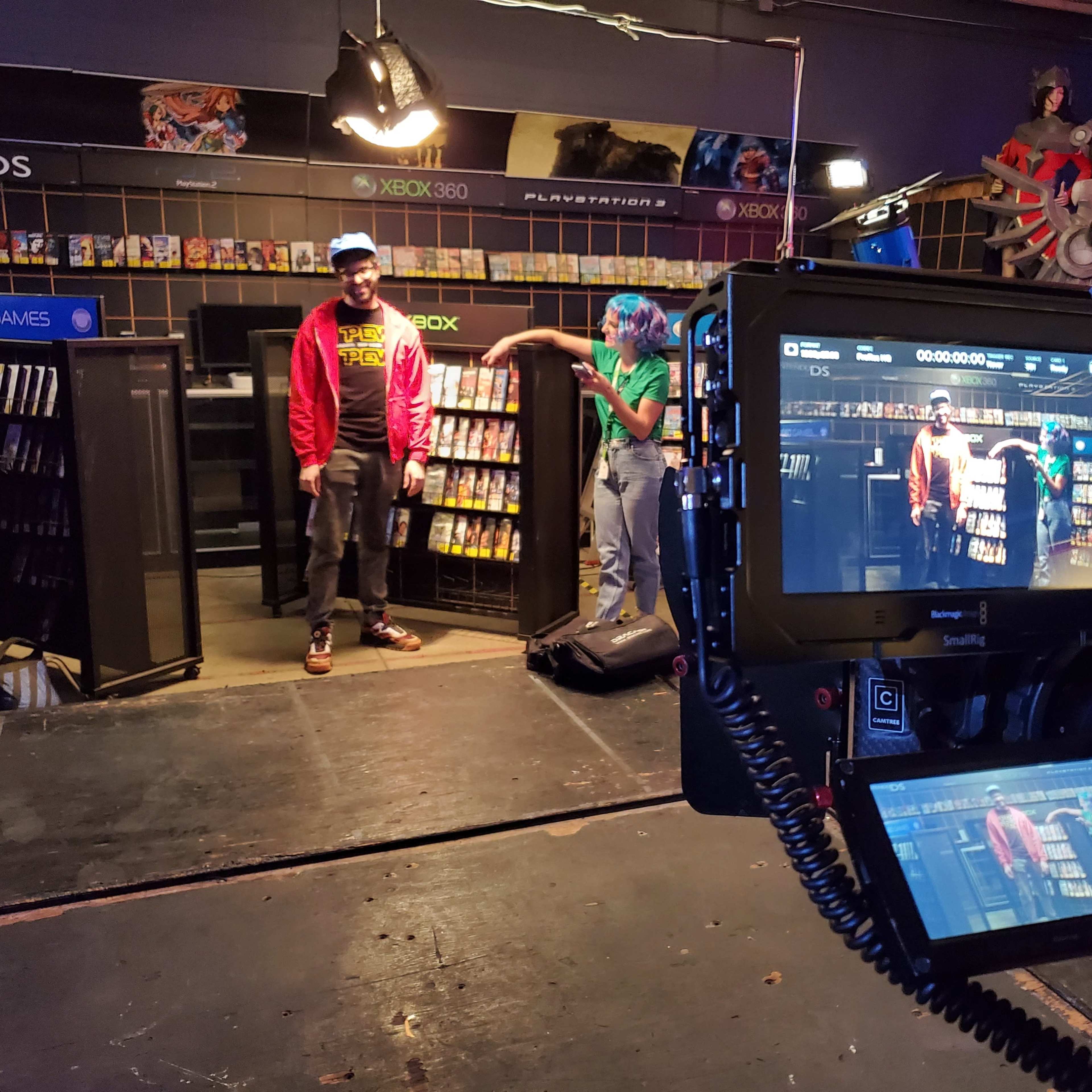 A film set featuring two people standing in a video game store aisle, with shelves filled with video games and a camera focused on them.