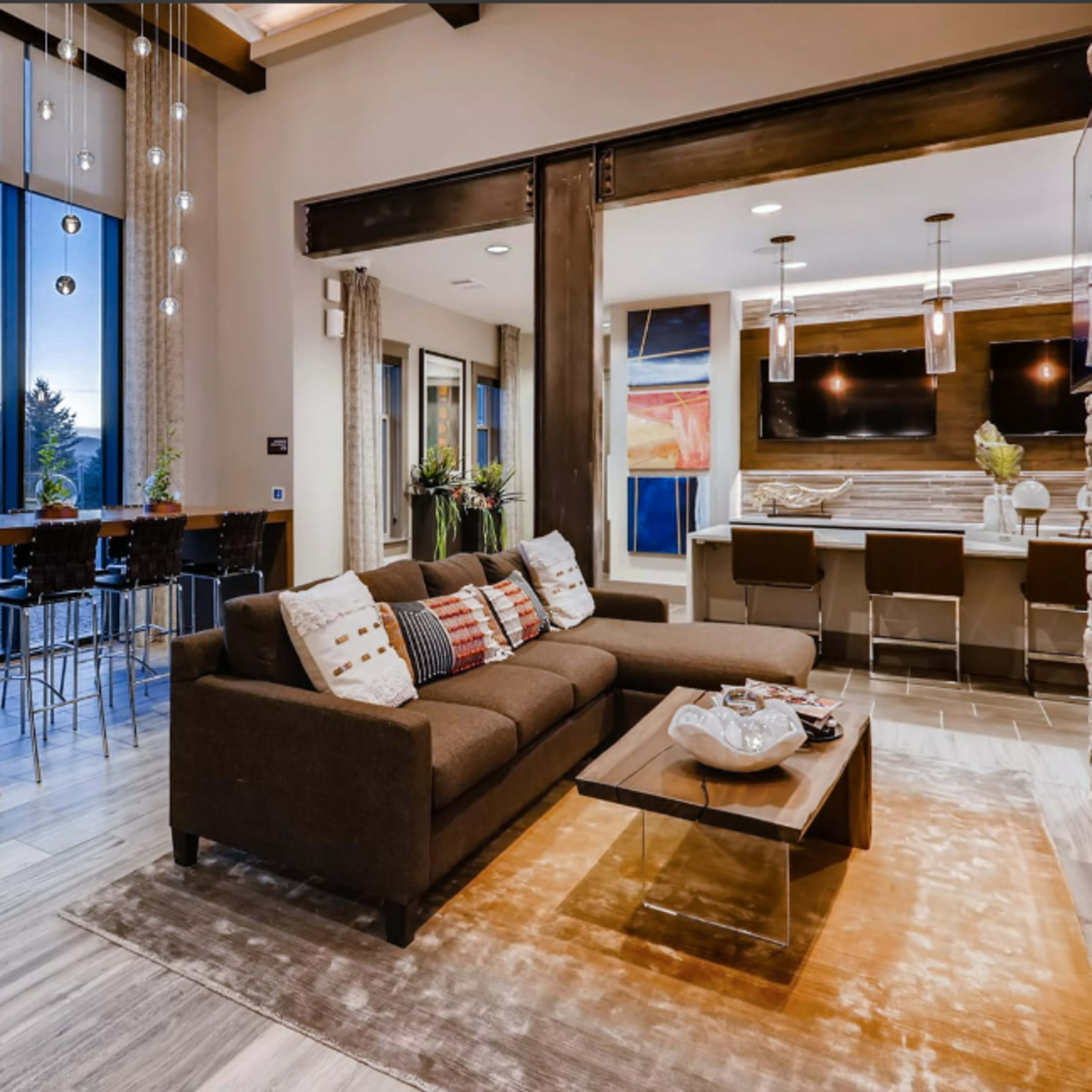 A stylish living area featuring a brown sofa, a coffee table, and a dining space with bar stools, all illuminated by pendant lights and large windows offering a view of the outdoors.