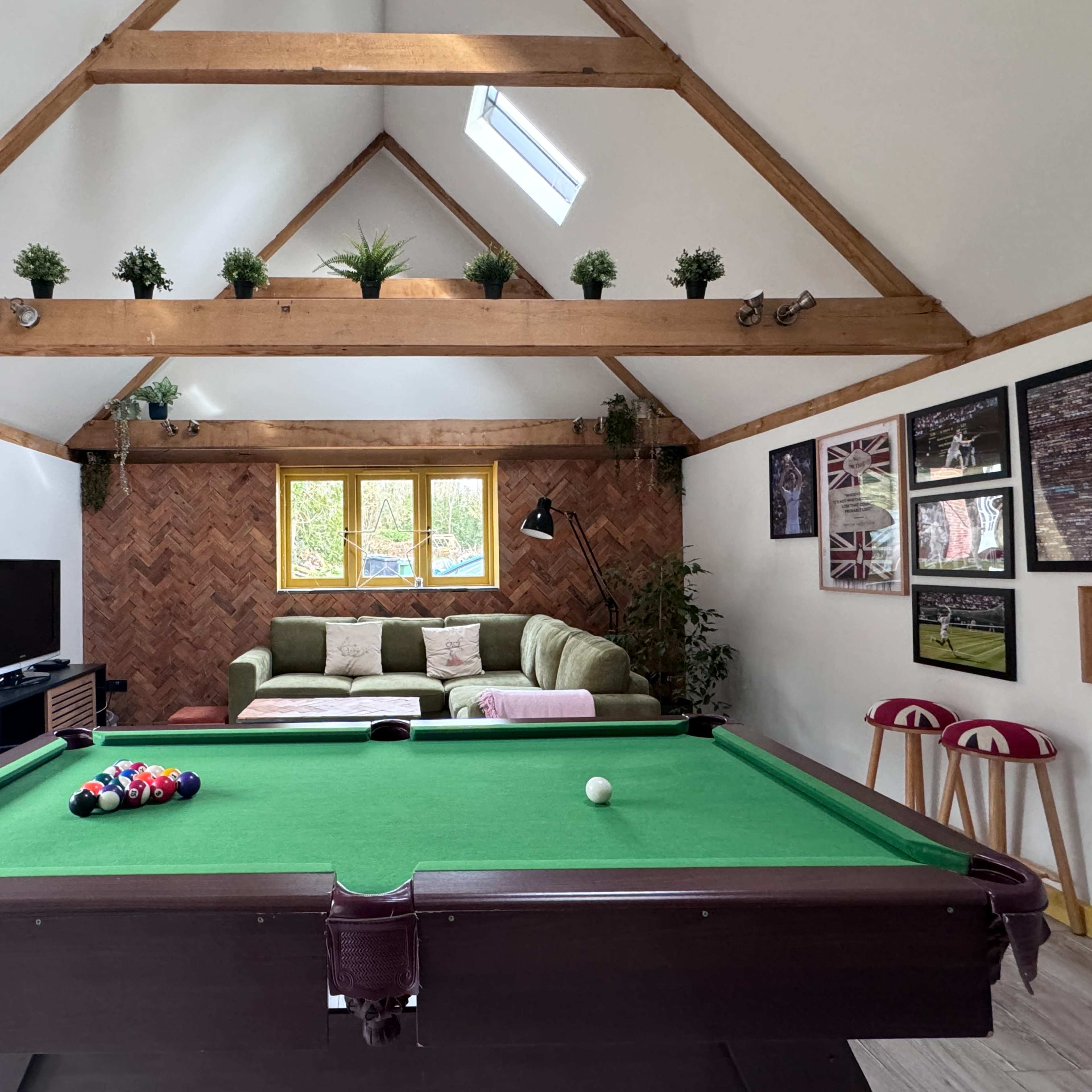 A games room with a green pool table in the foreground, a brown herringbone wall, and a seating area with a green sofa and plants against a white wall.