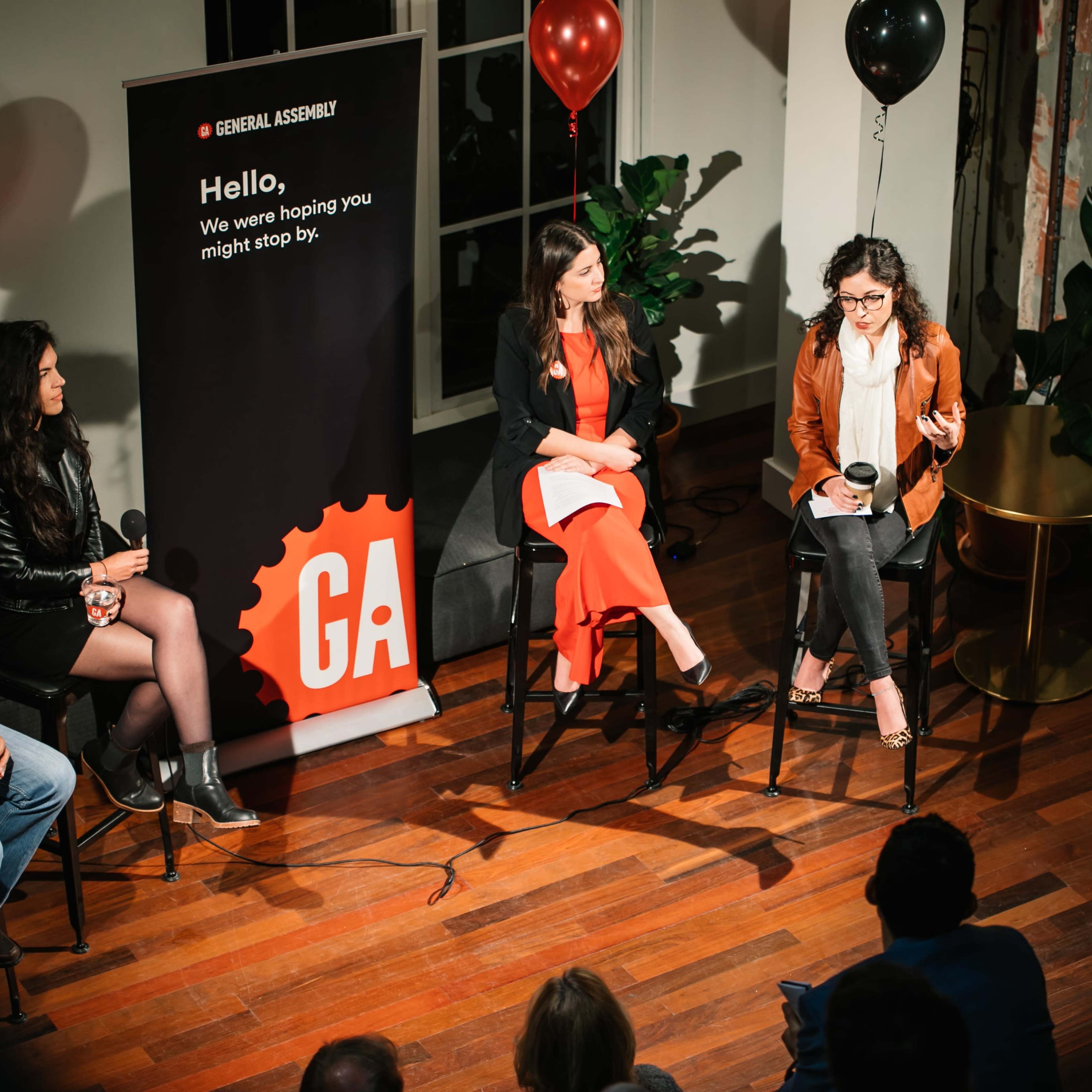 A panel discussion takes place in a venue featuring four speakers seated on chairs, with a microphone and a backdrop promoting General Assembly.