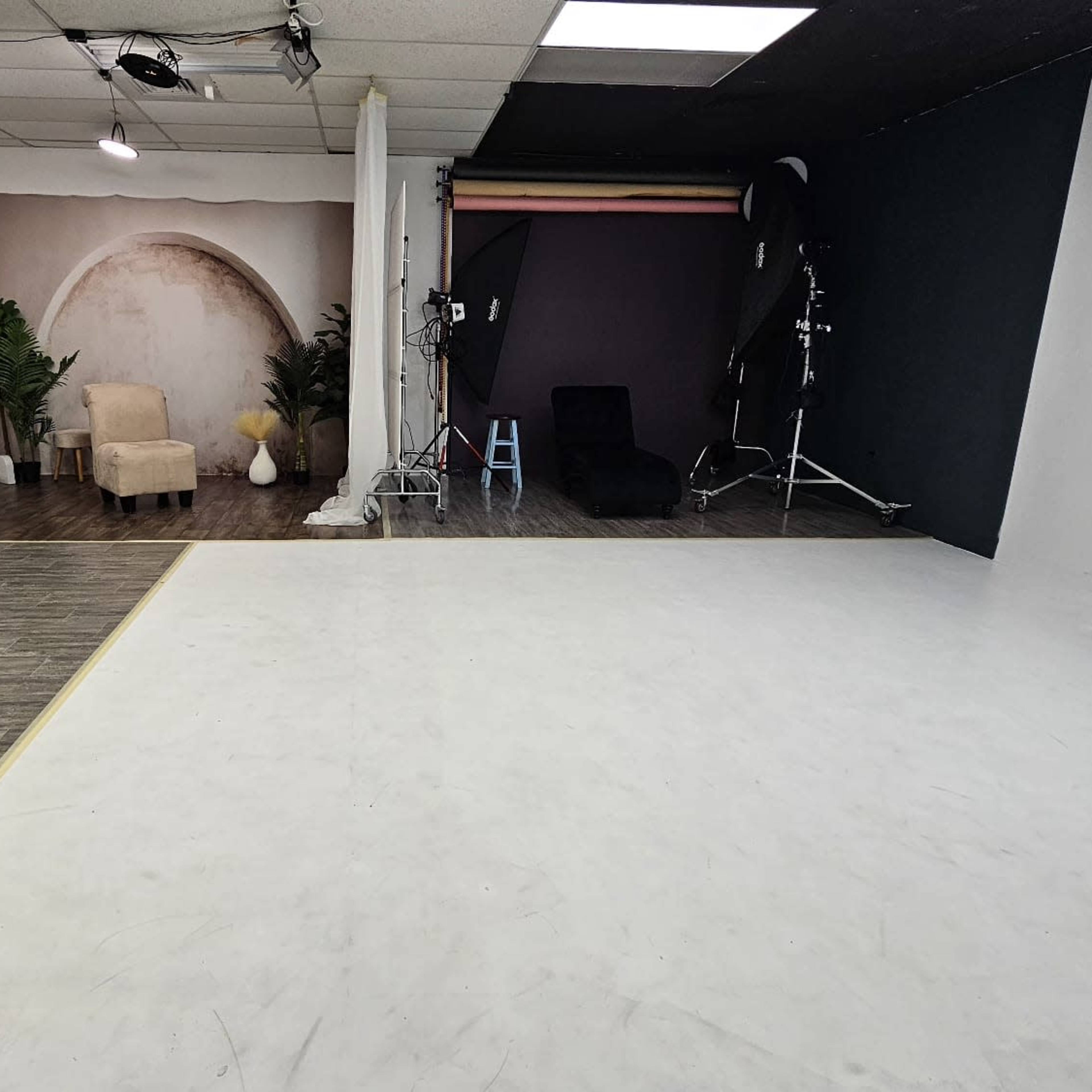 A spacious photography studio with a divided floor; one side features a white surface and the other has a wooden floor, along with a backdrop, chairs, and lighting equipment.