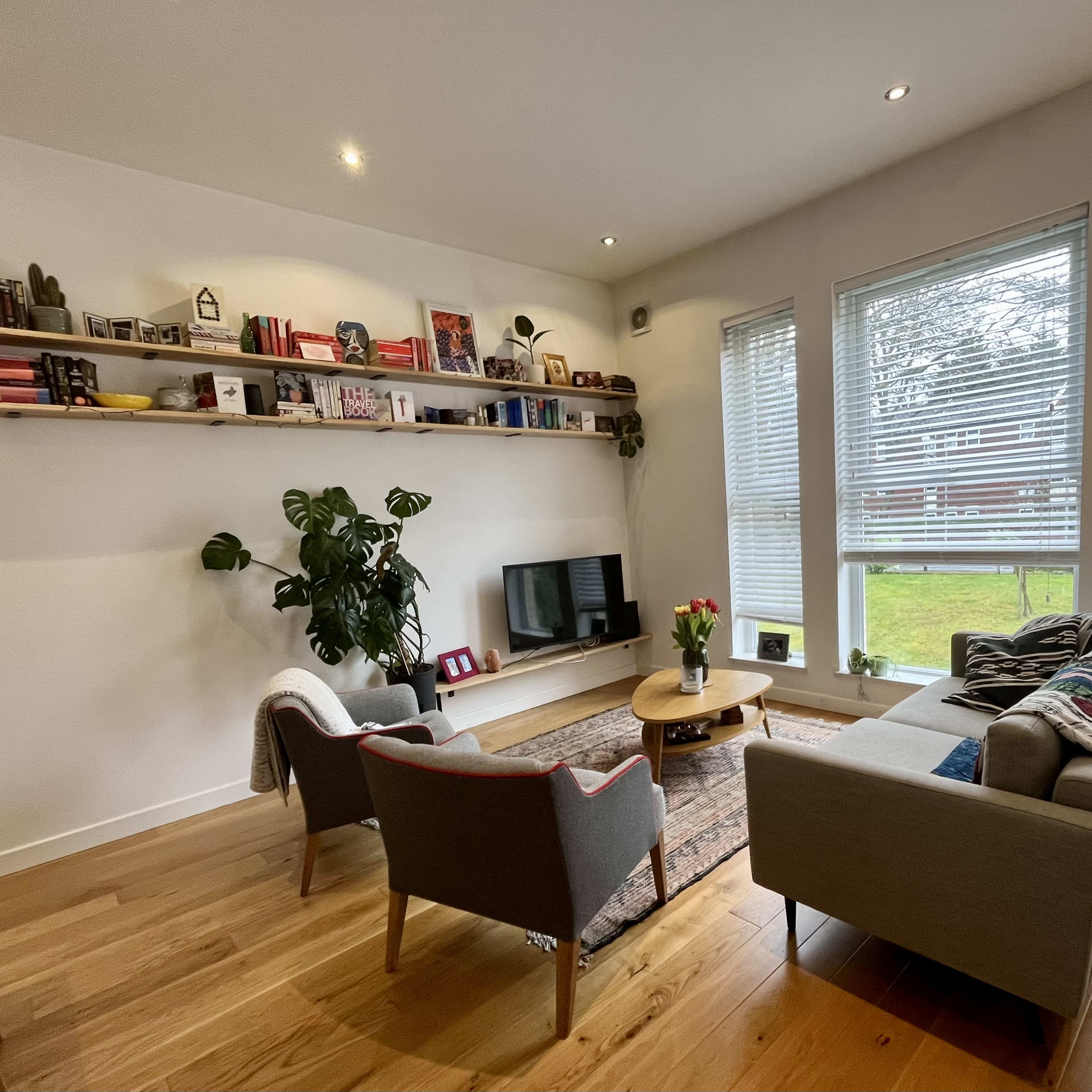A cozy living room features a gray sofa, two armchairs, a wooden coffee table, and a long shelf adorned with books and decor.