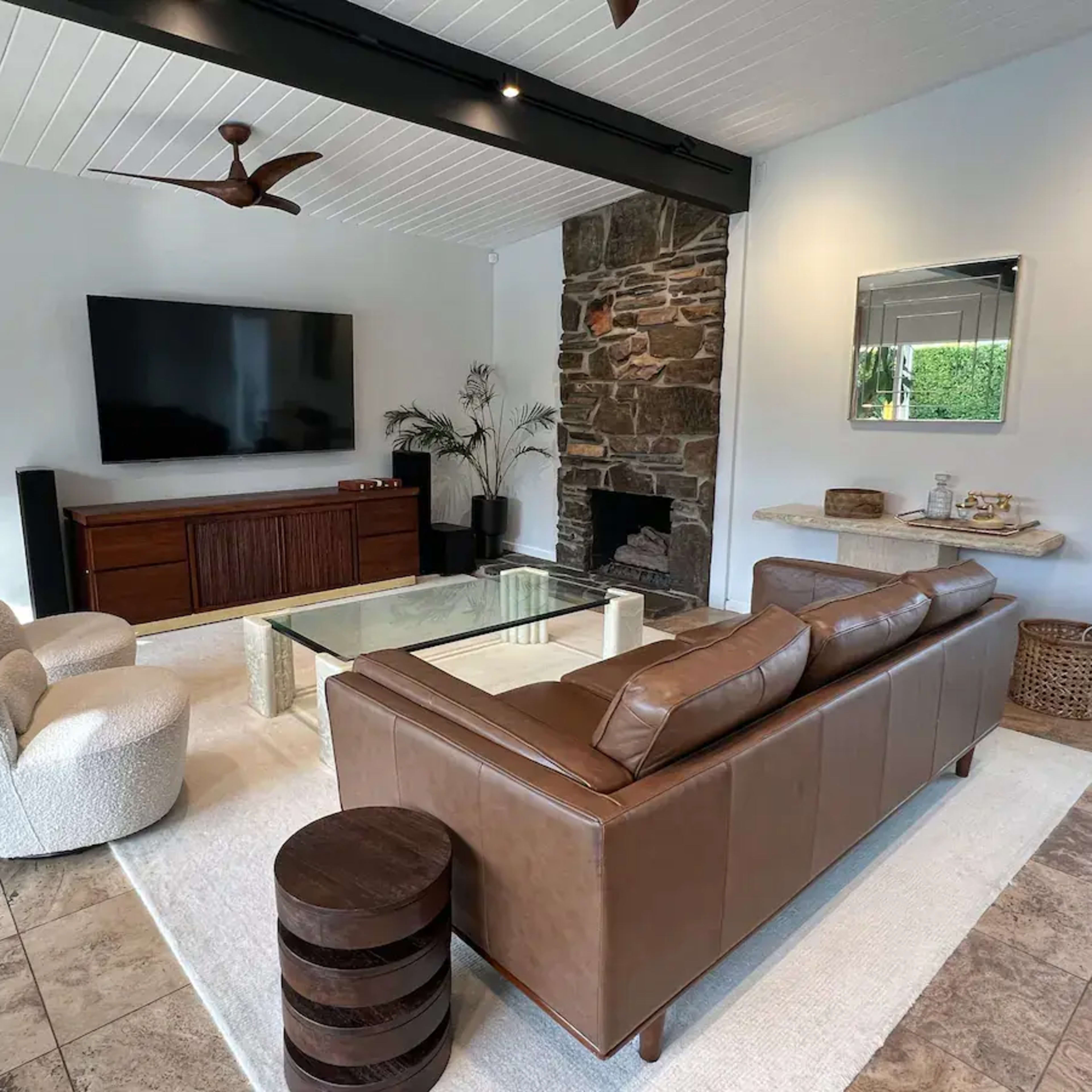 A modern living room featuring a brown leather sofa, a glass coffee table, a stone fireplace, and a wall-mounted television.