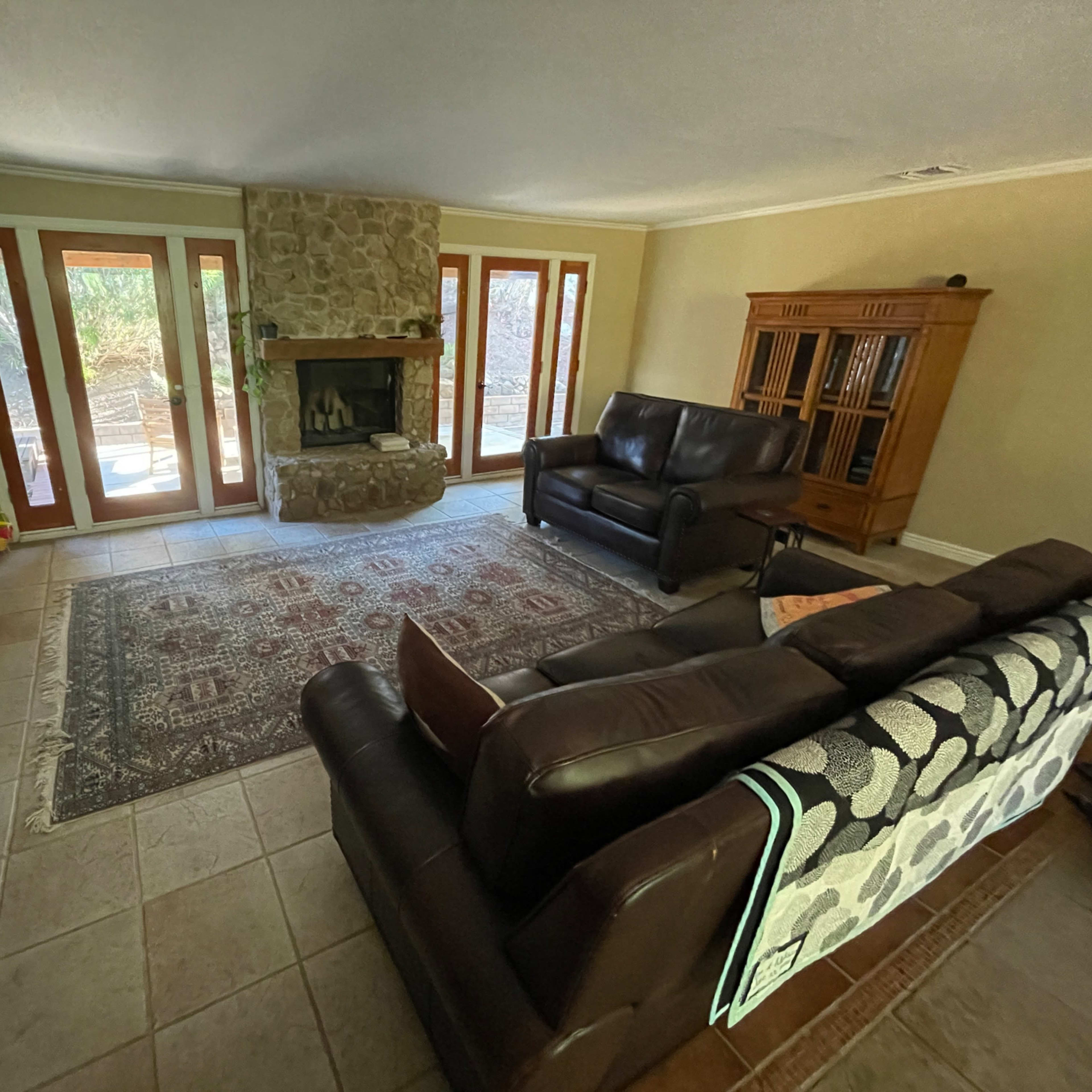 The living room features two leather sofas facing a stone fireplace, with large windows and a wooden cabinet along one wall.
