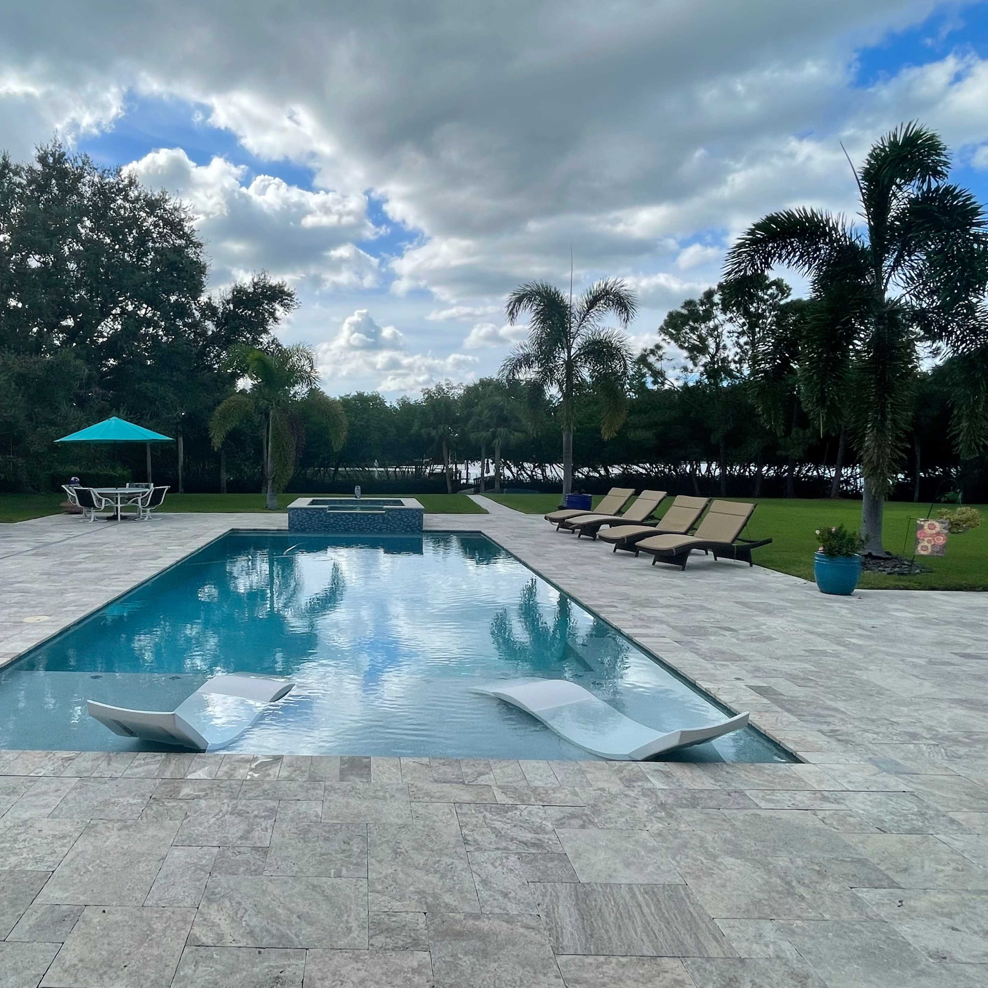 A rectangular swimming pool with lounge chairs and an umbrella is surrounded by a patio area and lush greenery under a partly cloudy sky.