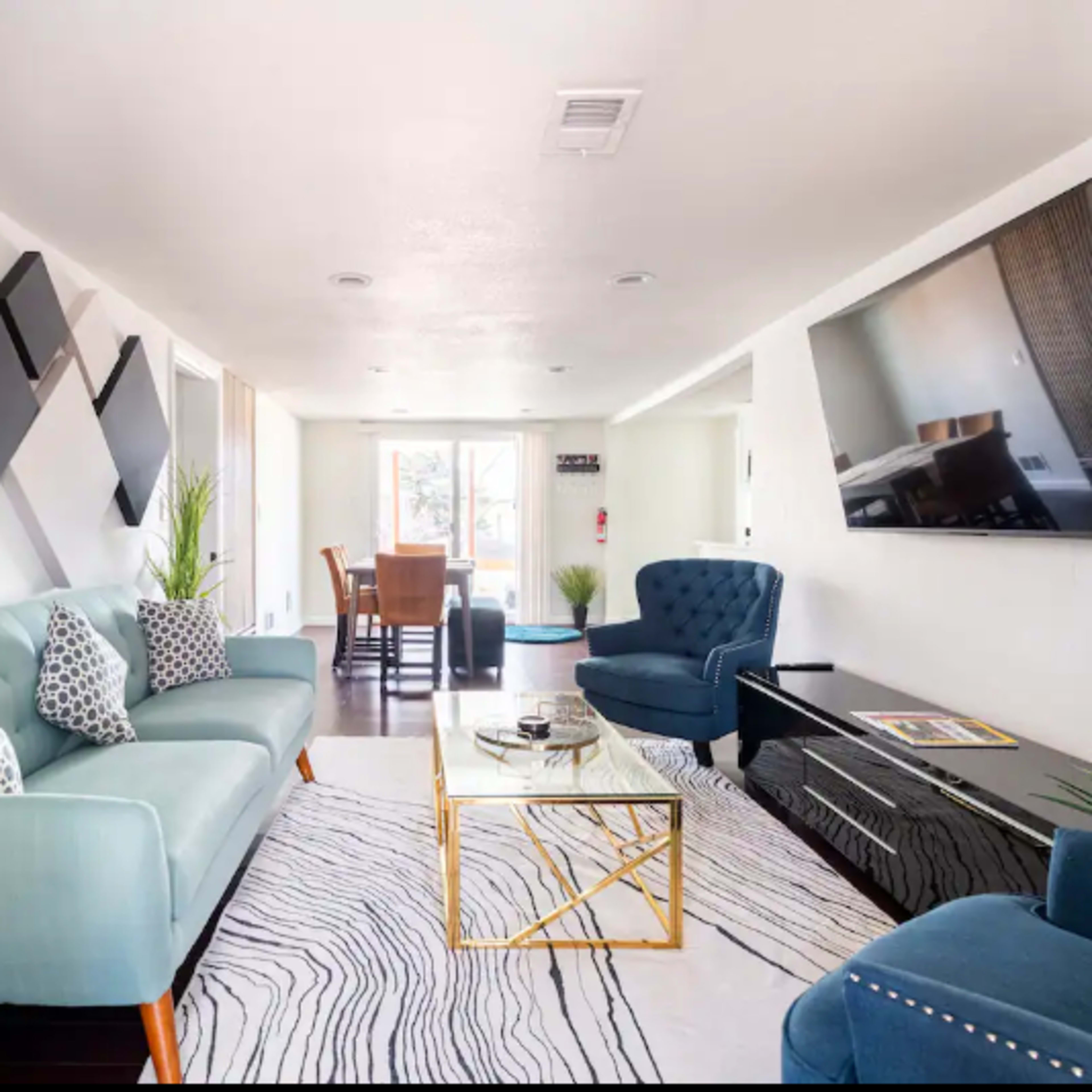 A modern living room features a blue sofa, two blue chairs, a glass coffee table, and a dining area in the background.