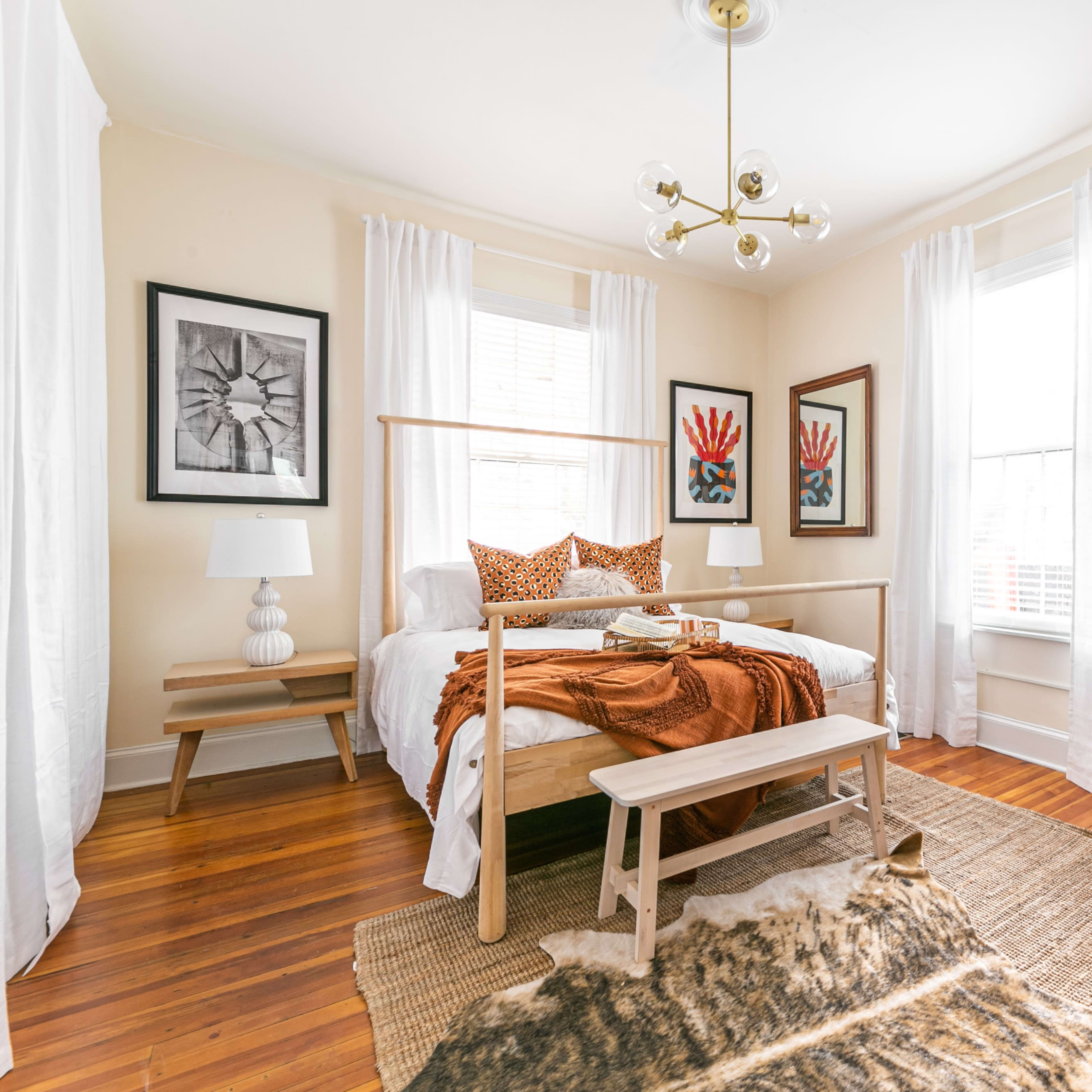 The room features a bed with a wooden frame, a desk, and decorative art on the walls, complemented by natural light from large windows.