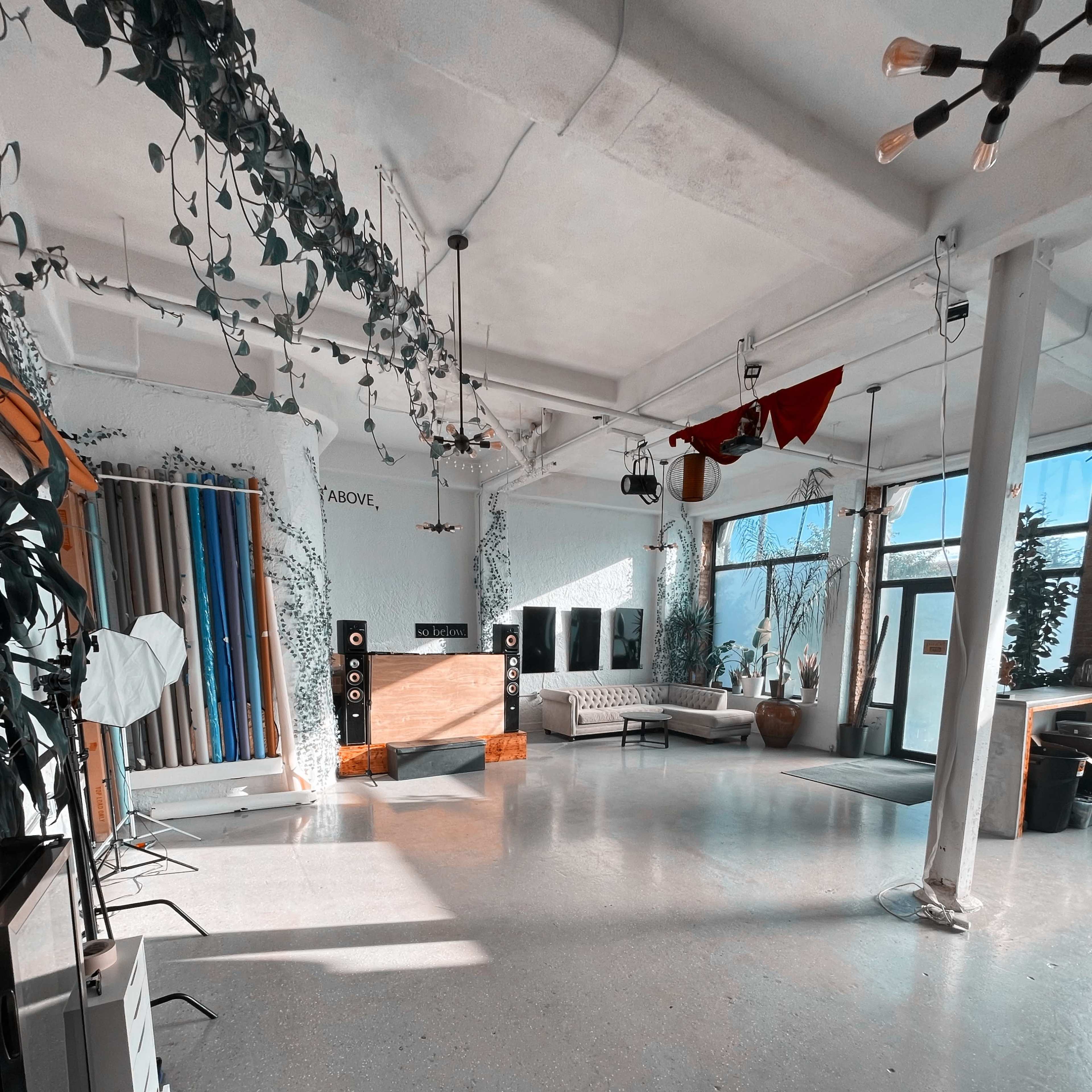 A spacious interior with large windows, green plants, a sofa, and audio equipment arranged against one wall.
