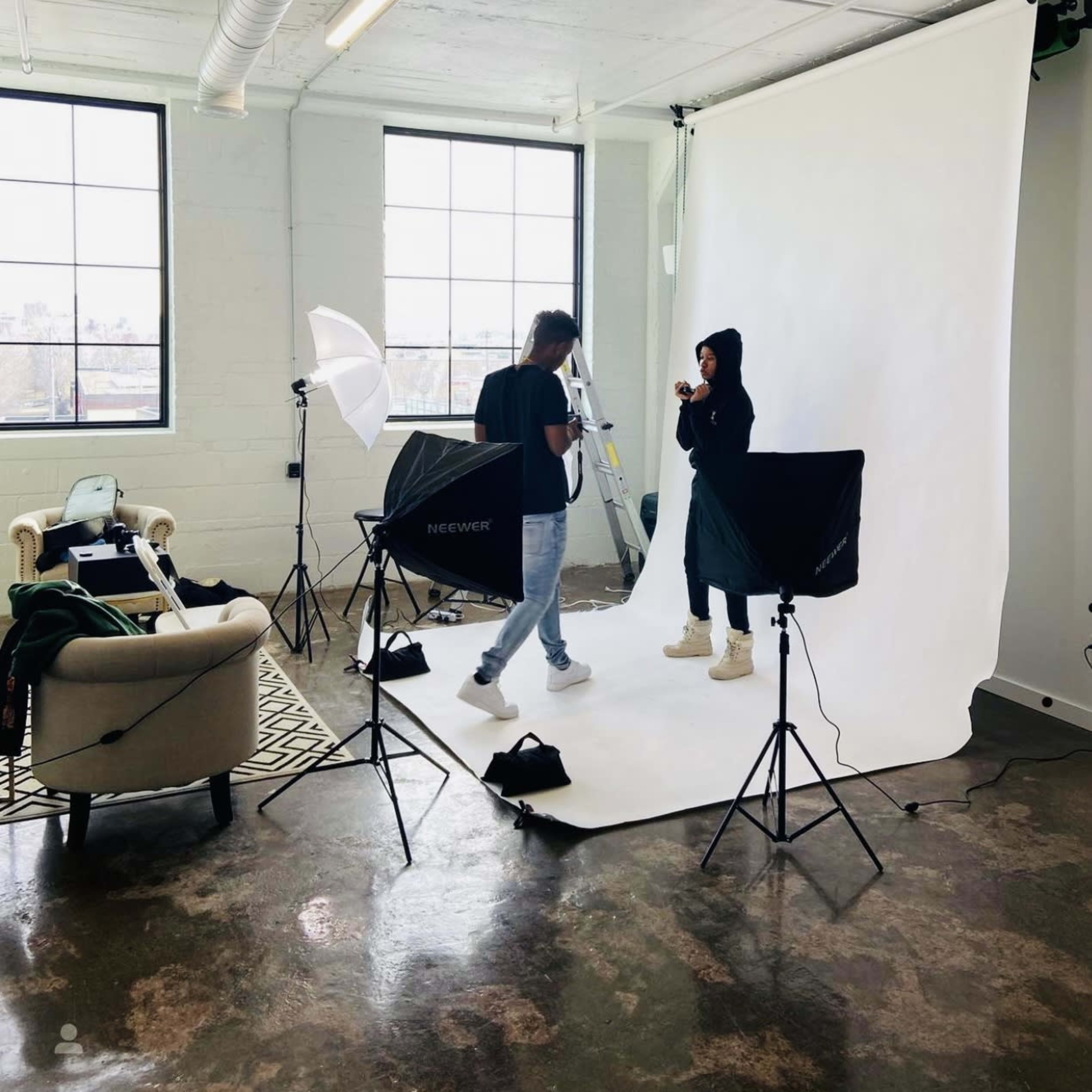 A photography studio features a model and photographer preparing for a shoot with lighting equipment and a backdrop.