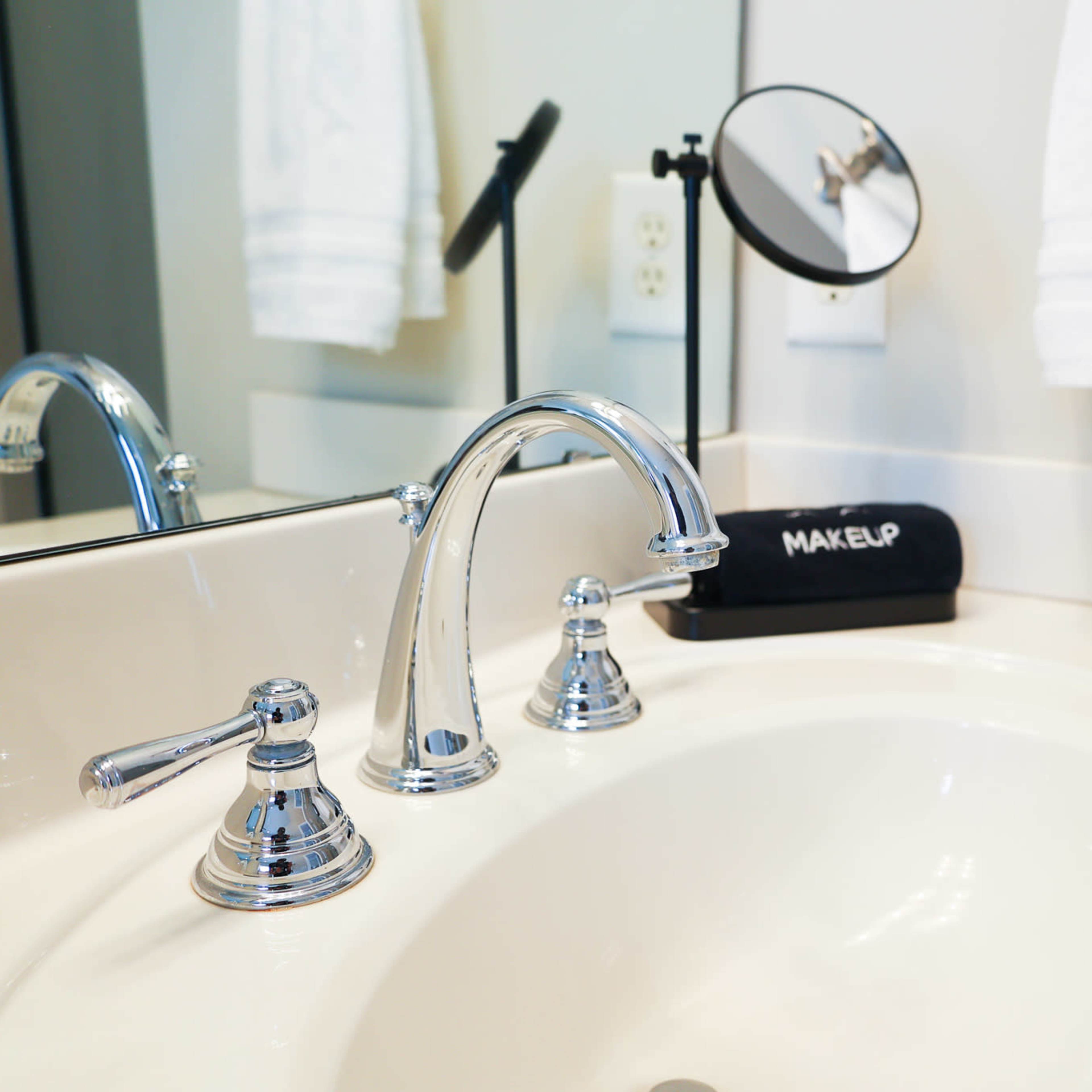 A bathroom sink with chrome faucets, a round mirror, and a black makeup organizer sits on a countertop.
