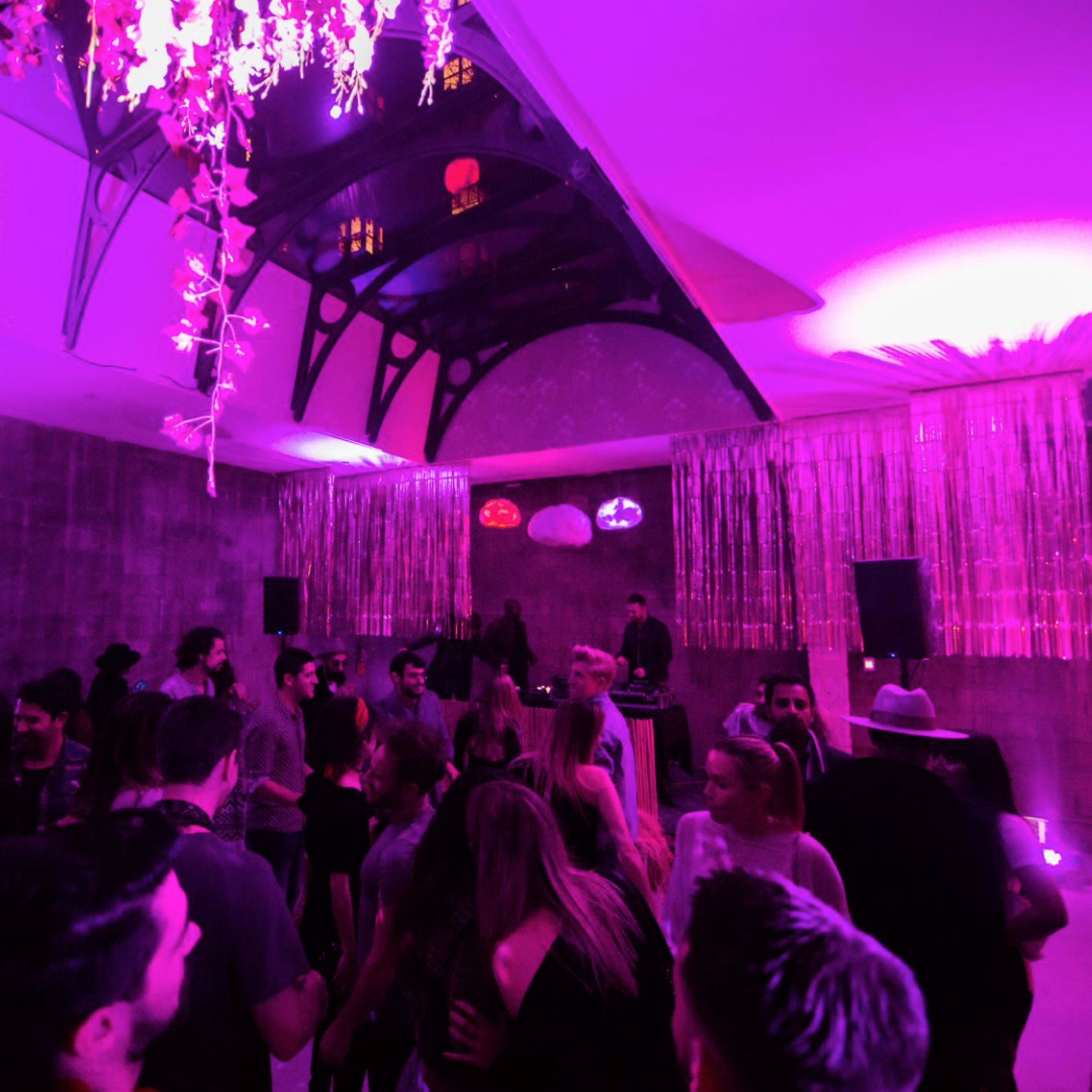 A crowded indoor venue is illuminated by purple lighting, featuring a DJ setup and decorative elements like hanging flowers and glittering streamers.