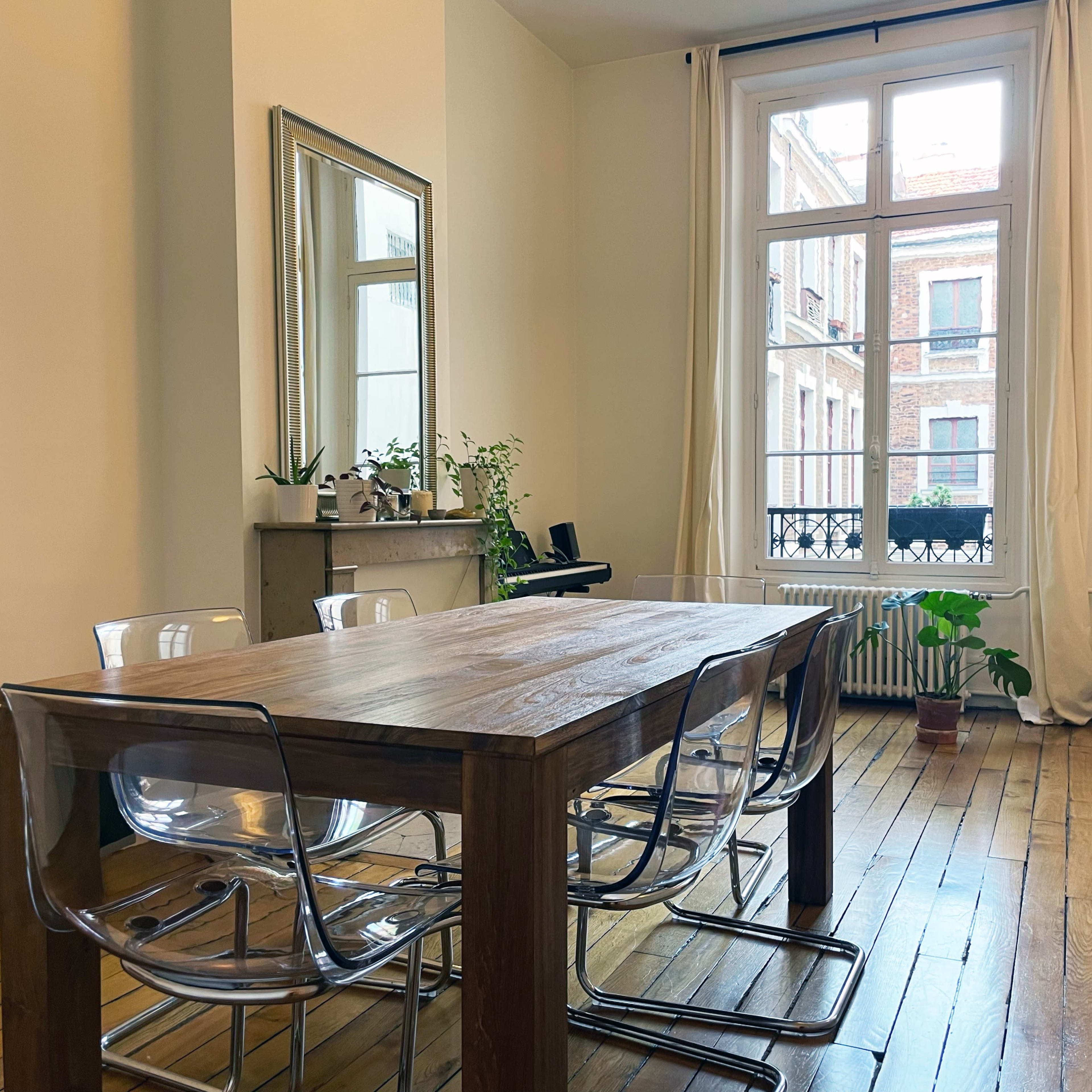 A wooden dining table with clear chairs is positioned in a room that features a large window overlooking a street, with plants and a guitar in the background.