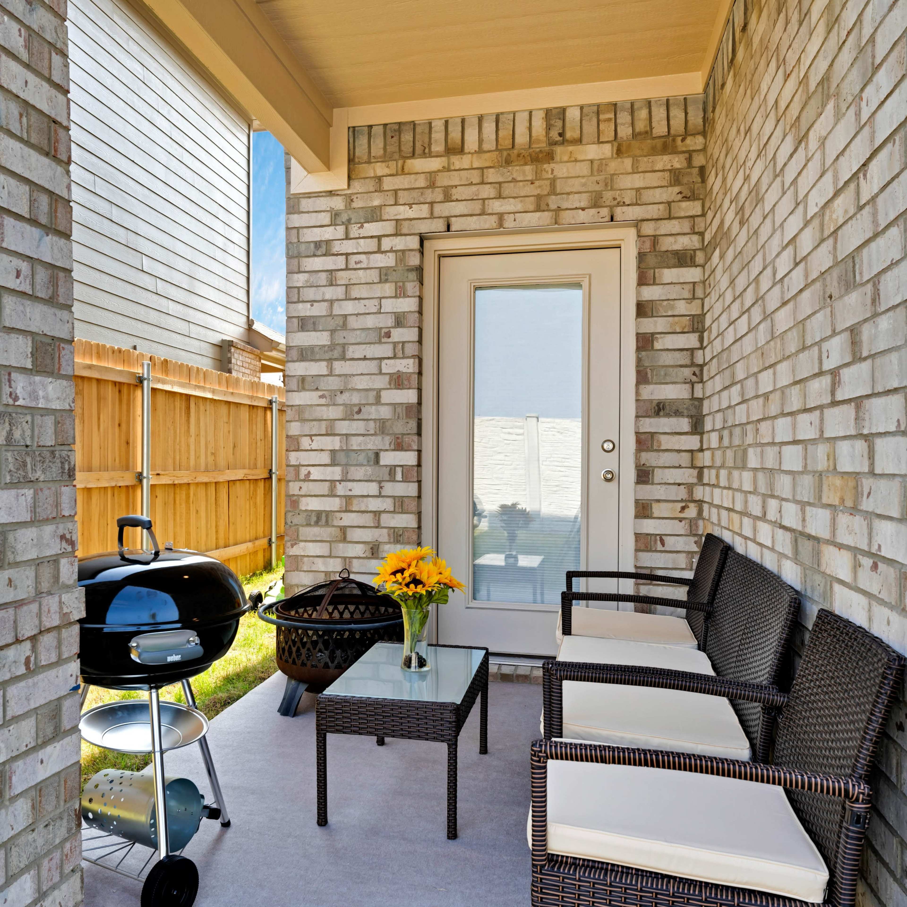 A covered patio area with a small table, chairs, a grill, and a flower vase in a residential setting.