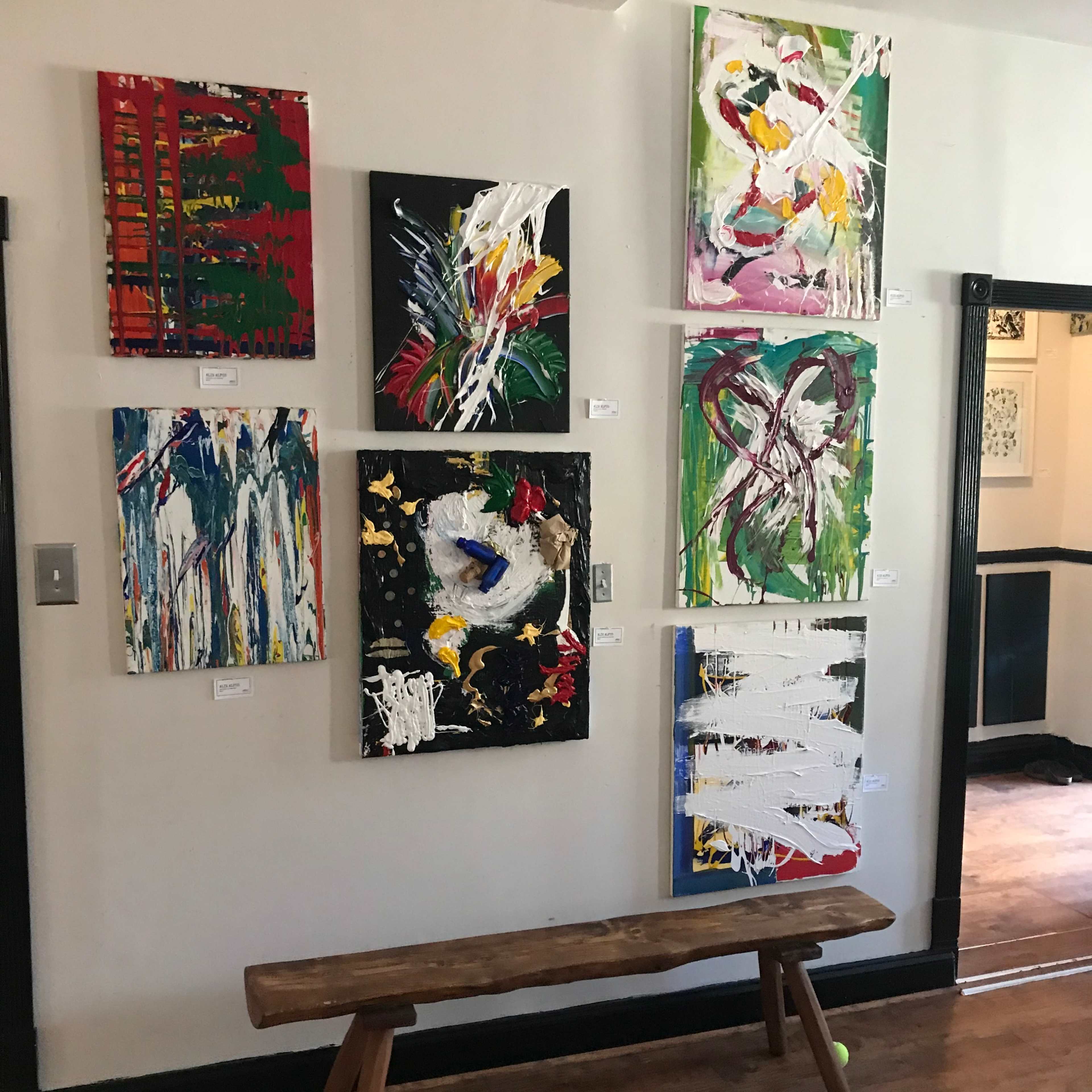 A wooden bench sits in front of a wall featuring eight abstract paintings in various colors and styles.