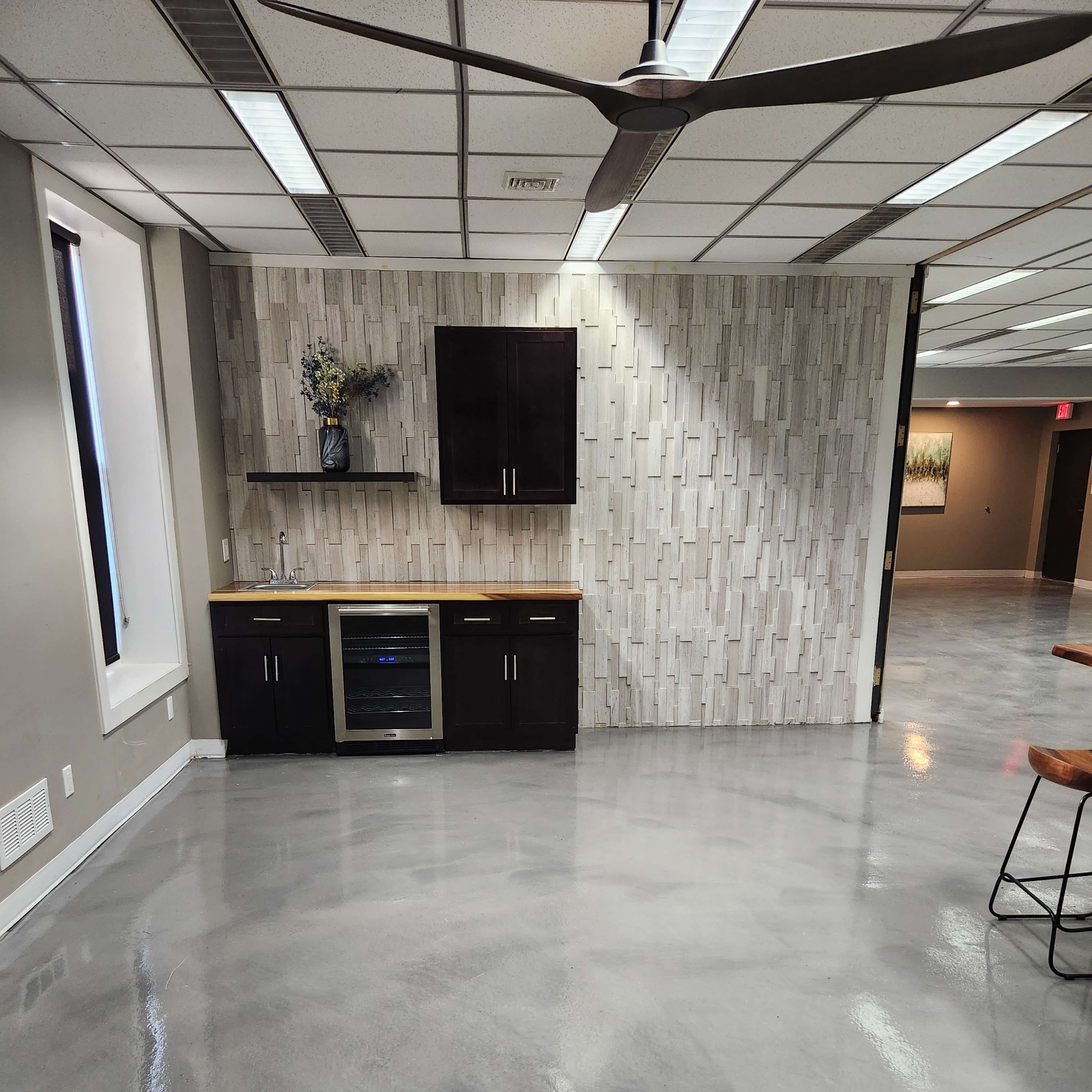 A modern kitchen area with a countertop, cabinetry, and a glass-front fridge, set against a textured wall in an open space with polished concrete flooring.
