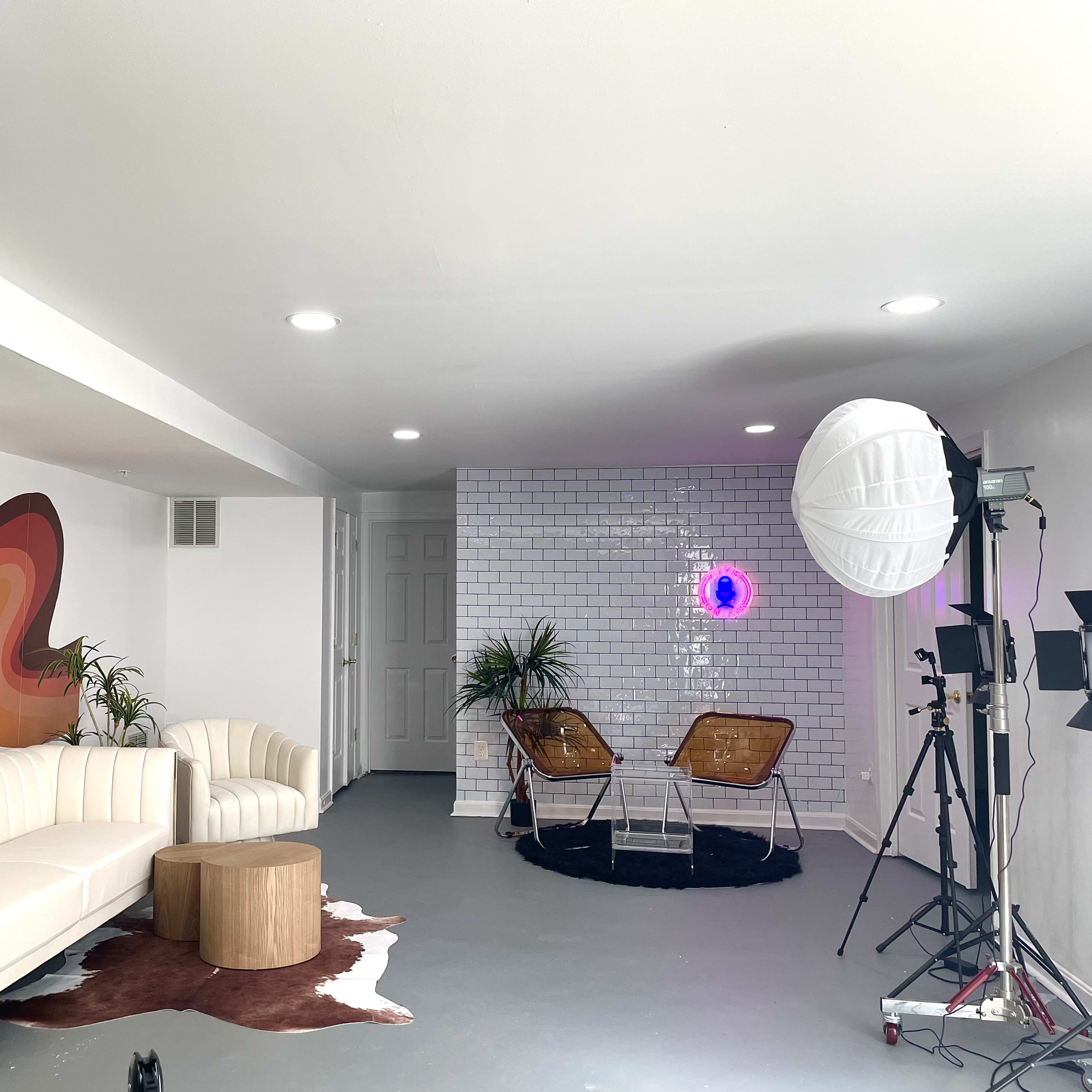 A modern indoor studio space featuring a white sectional sofa, a wooden coffee table, two stylish chairs, a circular light fixture, and a textured wall with purple lighting.