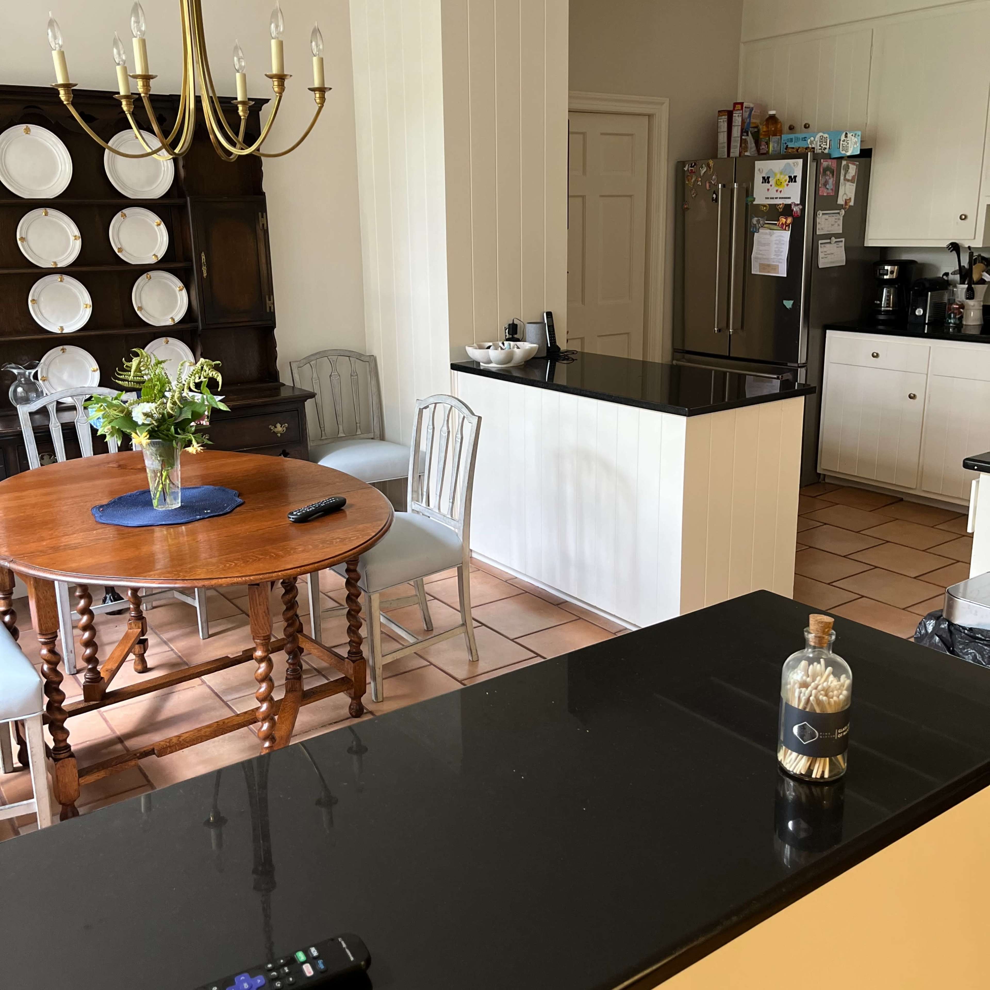 A kitchen and dining area features a round wooden table with chairs, a black countertop, and a chandelier above, with plates displayed on a nearby cabinet.