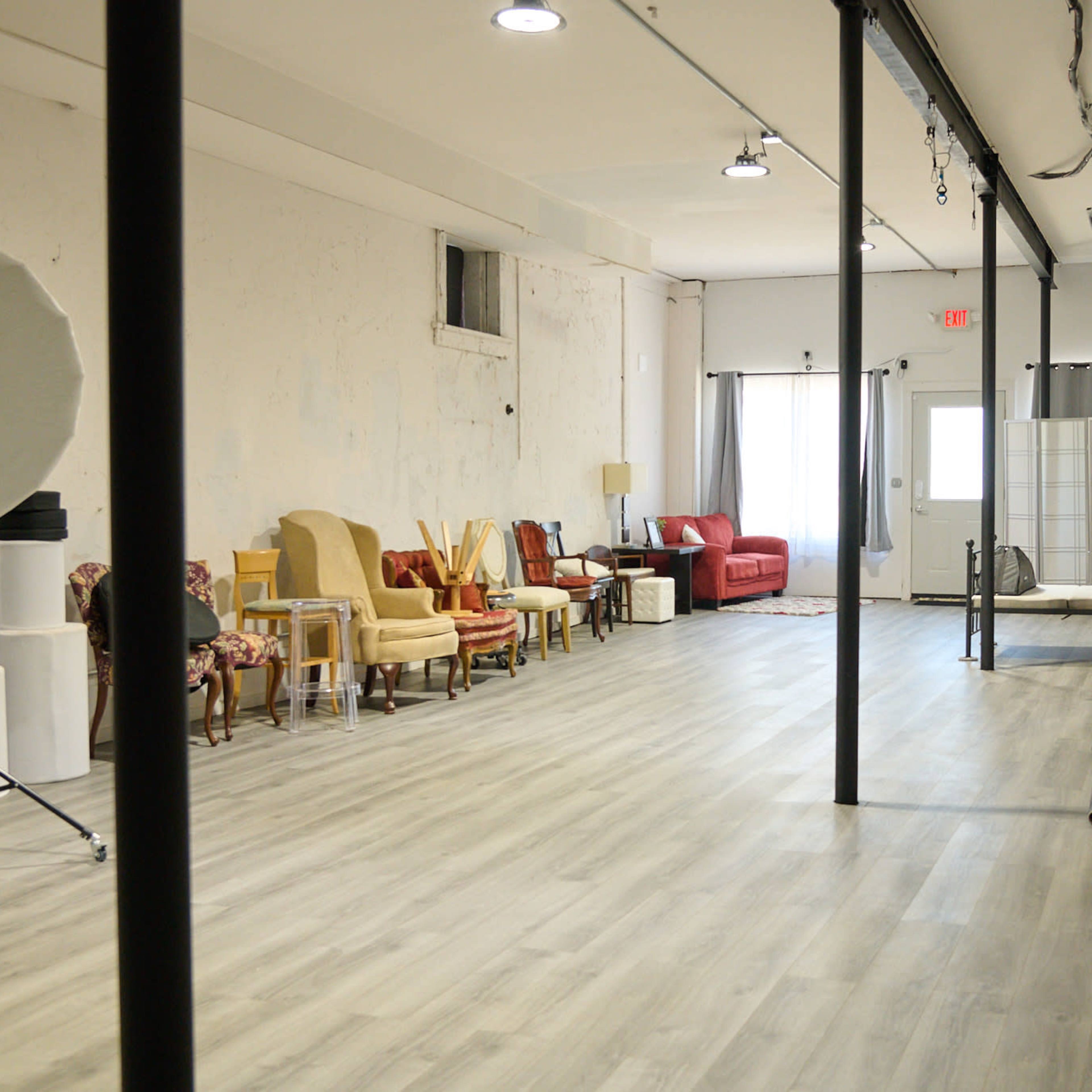 A spacious studio with various seating arrangements, including chairs and a sofa, along one side and a bed setup in the back corner.
