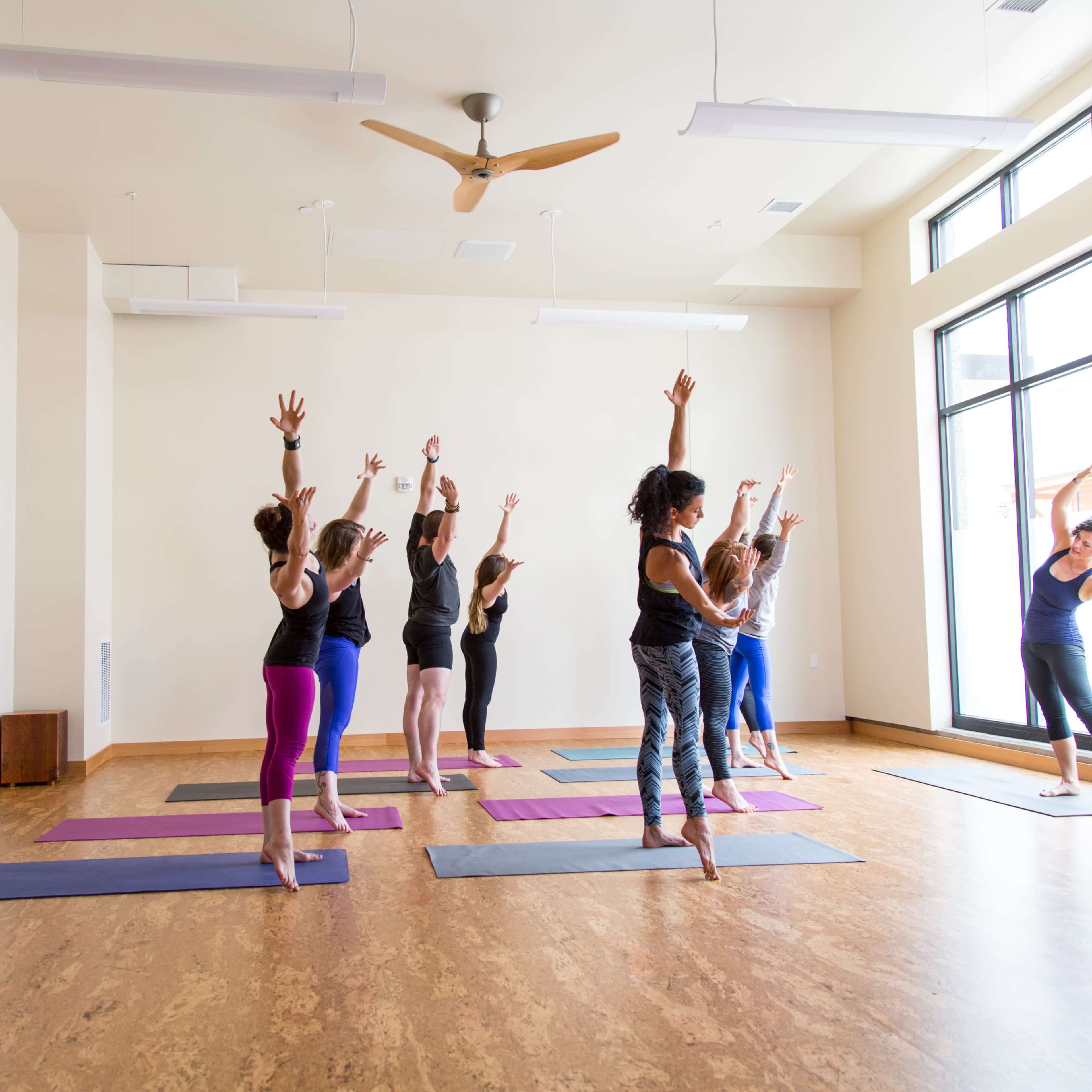 A group of people practices yoga in a spacious studio with large windows and wooden flooring.