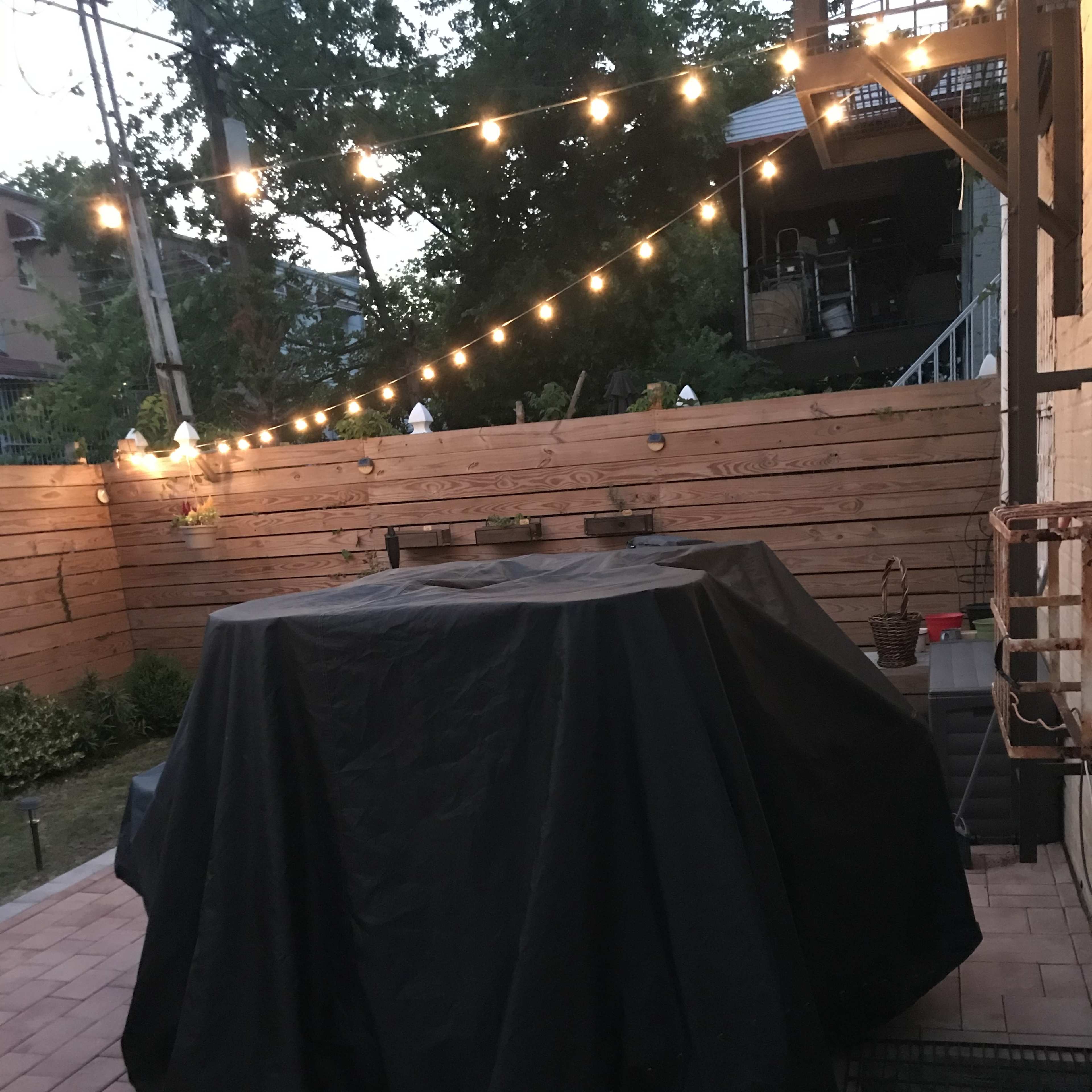A covered grill sits on a patio surrounded by string lights and wooden fencing.